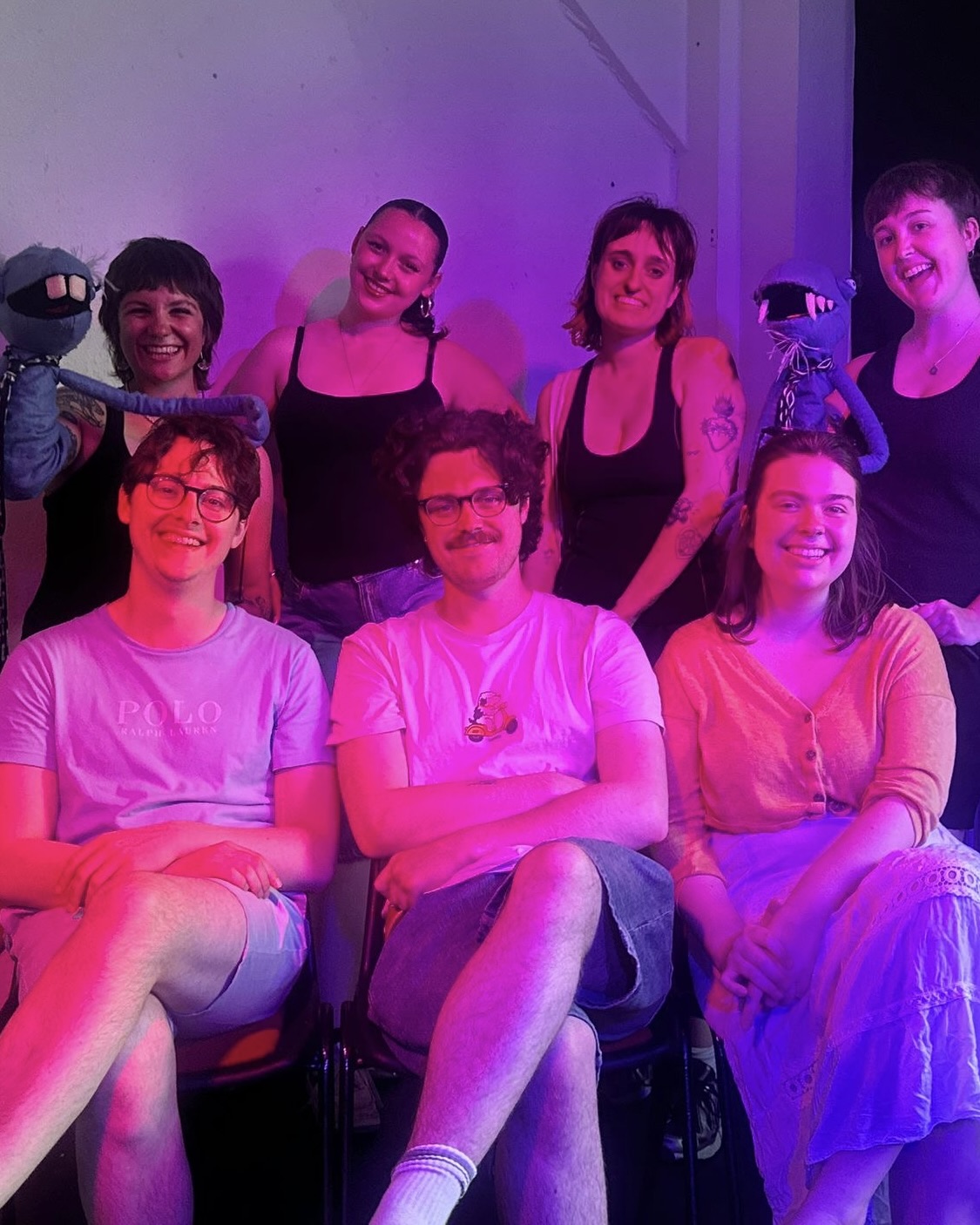 Group of seven people sat smiling under bright pink lighting, two on the top left and right holding blue puppets.