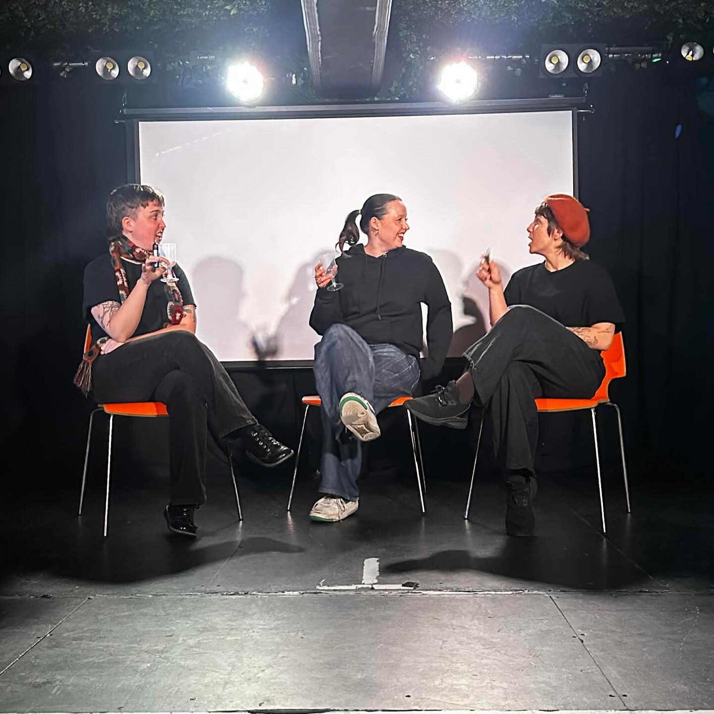 Three people sat on red chairs on a black stage space, lights and projection screen behind.