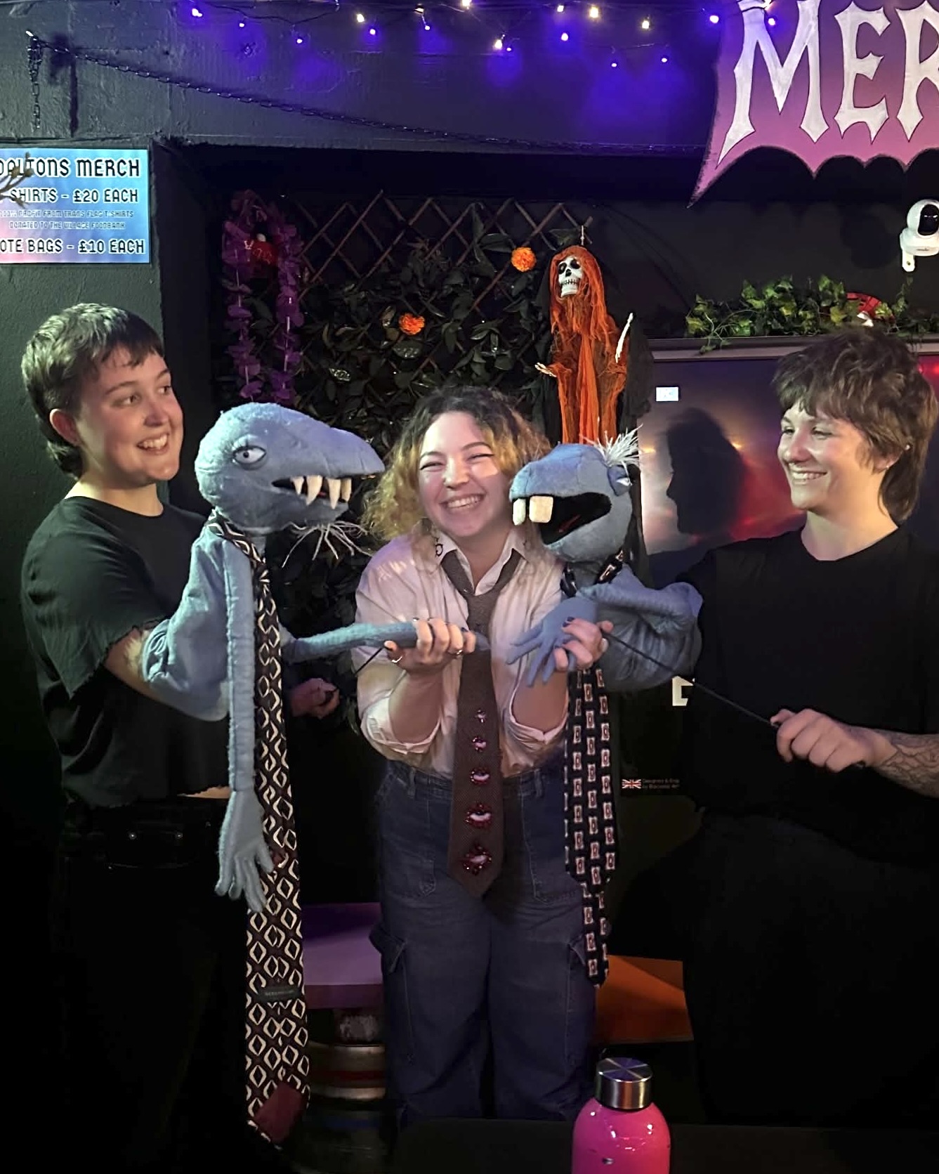 Three people stood smiling with blue puppets in a dark, indoor venue, purple lights above.