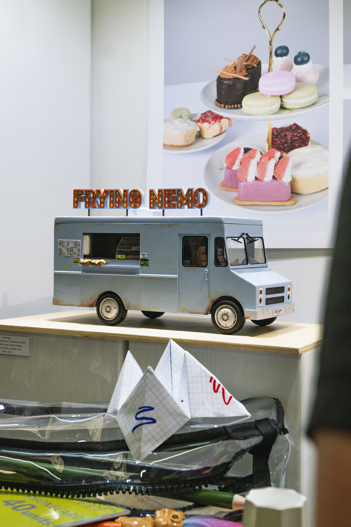 Model of a light blue van with red sign above that reads 'Frying Nemo', photo of model cakes in background.