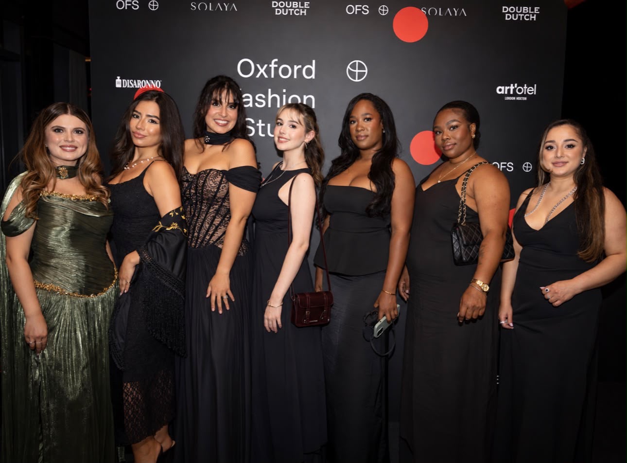 Seven women pose in evening dresses on a red carpet, in front of an Oxford Fashion Studio backdrop with sponsor logos.