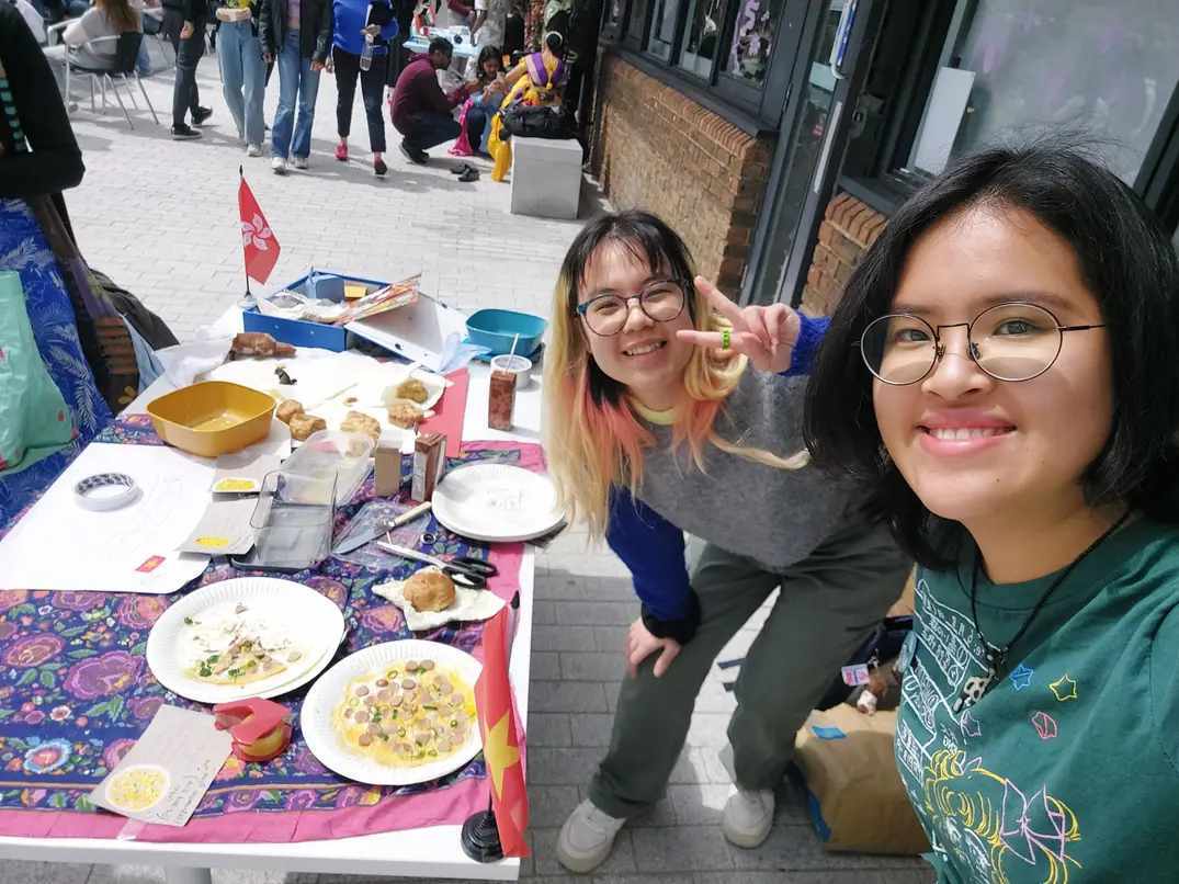Two girls in a selfie in the AUB courtyard at the international table with various food dishes.