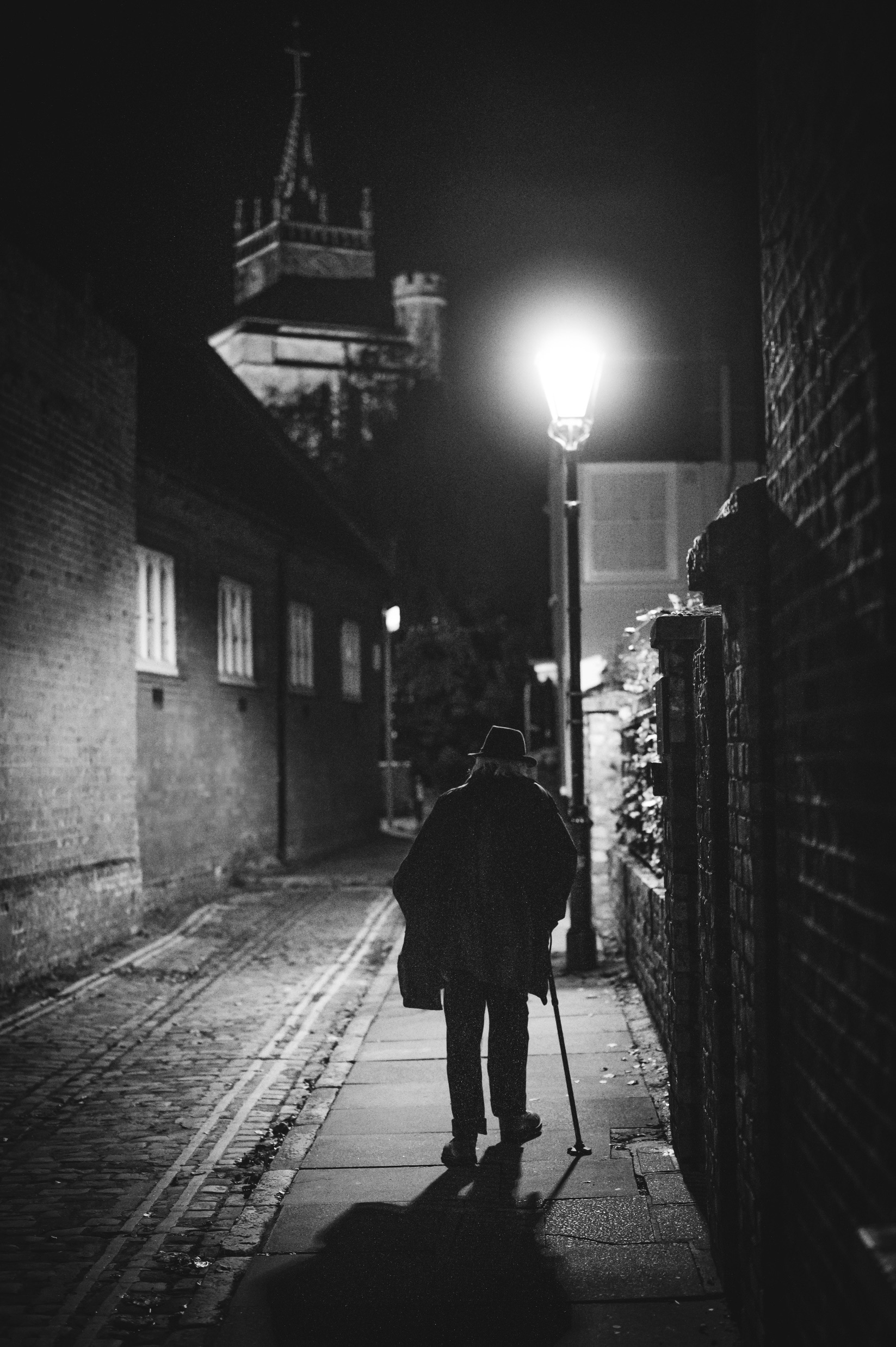 An elderly man walks down a cobblestone alley at night, using a cane. A street lamp illuminates the path, and a church spire is visible in the background, contrasting against the dark sky.