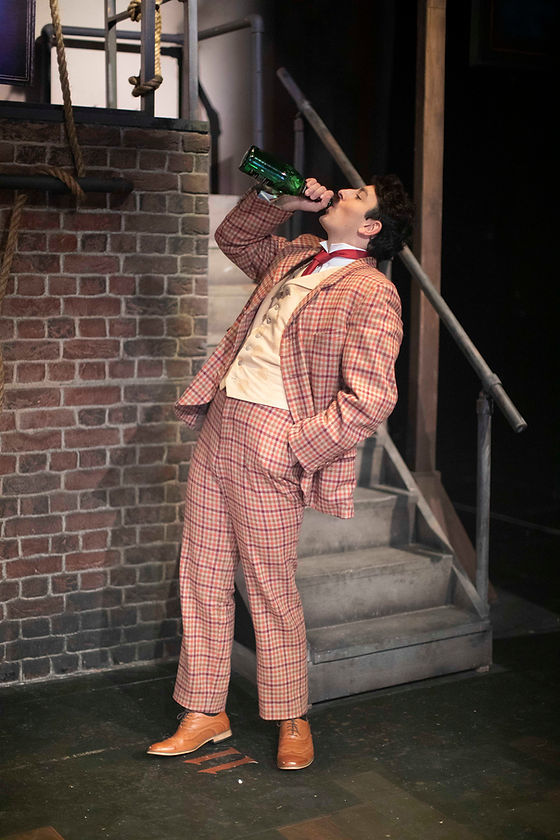 A man in 19th Century costume stands with one hand in a trouser pocket and swigs from a bottle.