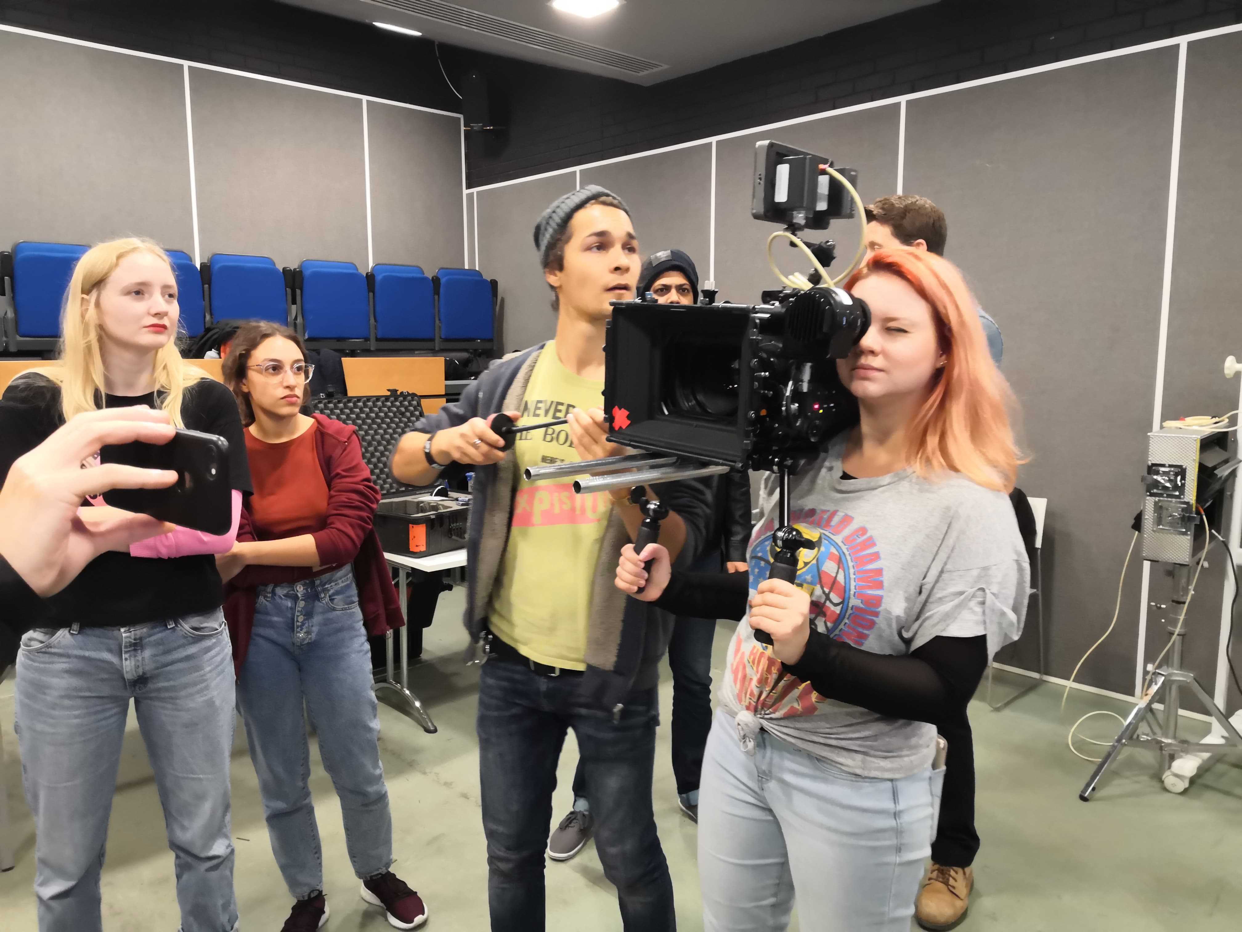 Person in a student holding a large camera with both hands and peering through it. Other people are to the side of them in the studio.