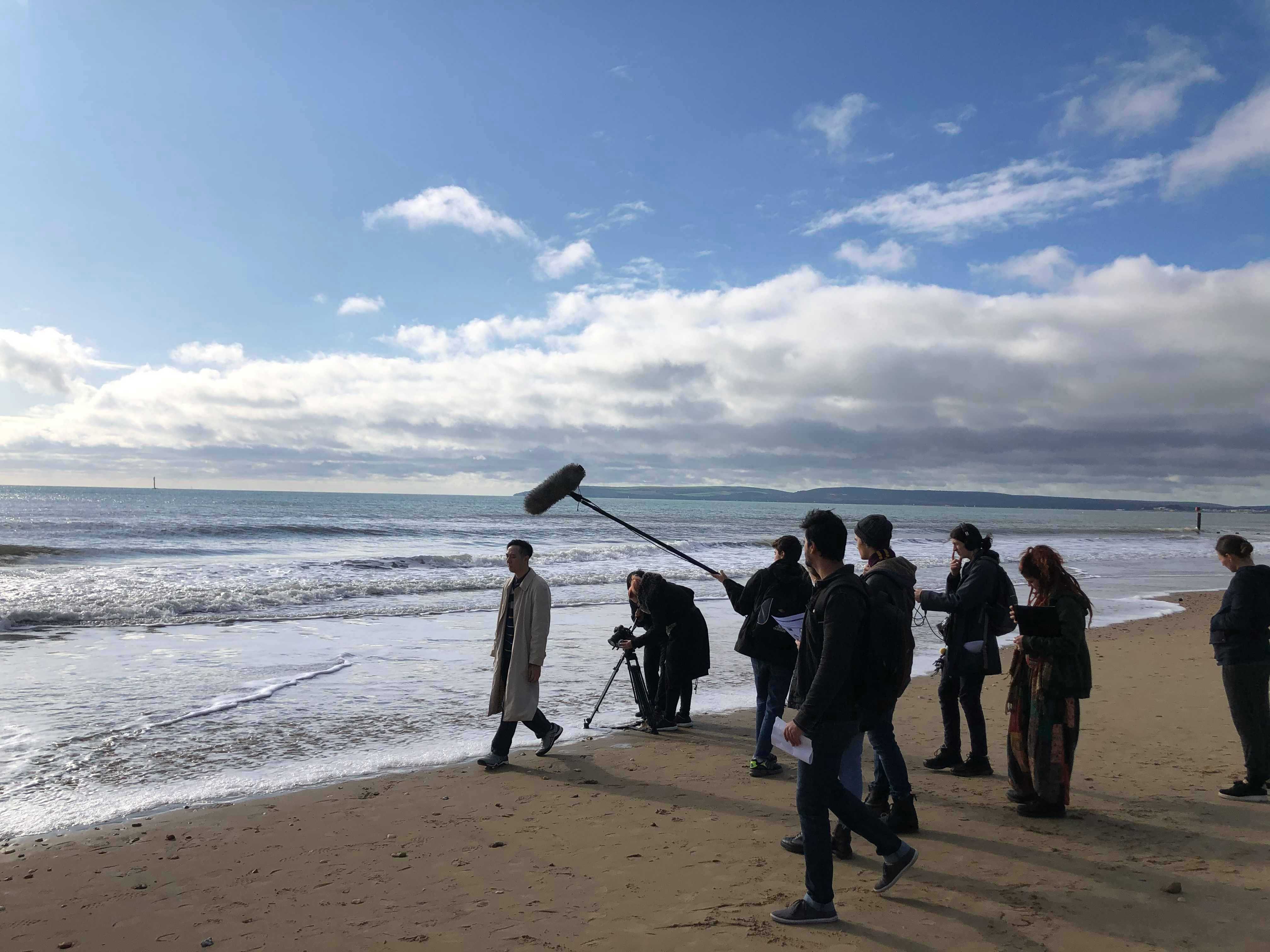 A film crew gathers on a beach and shoots a scene with an actor in costume walking along a shoreline.