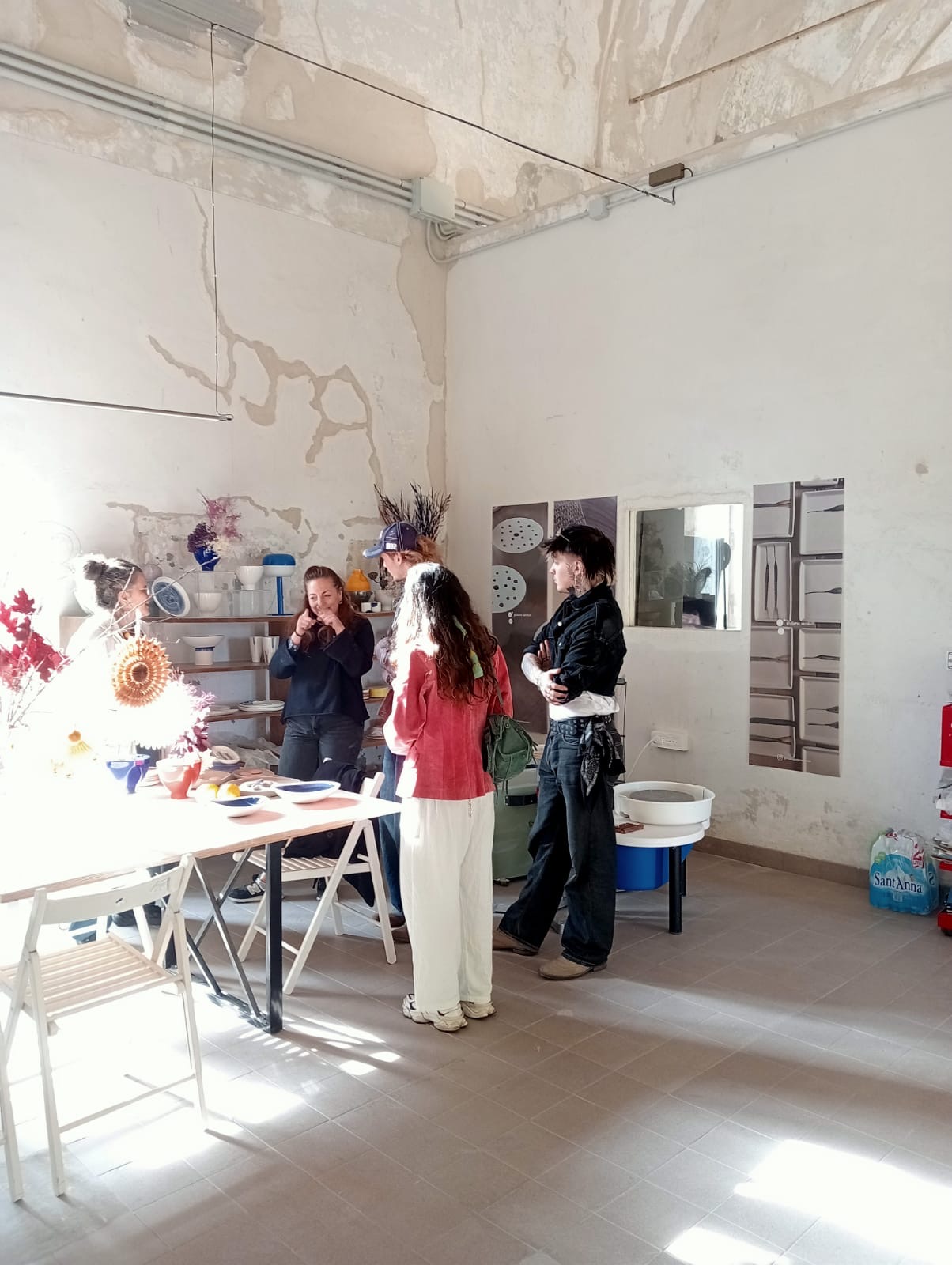 Three people stand in a bright studio beside a table of colourful paper flowers and craft materials, with peeling plaster walls and mirrors in the background.