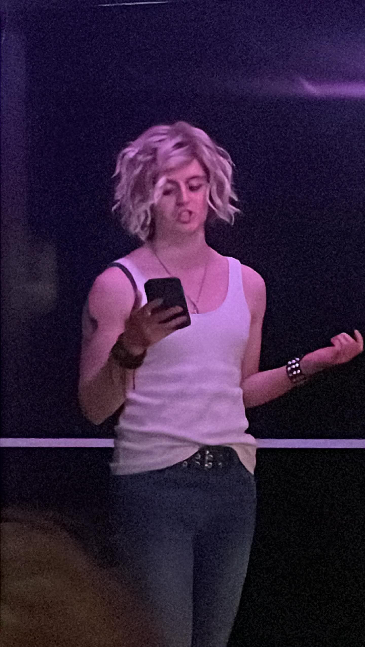 Person with short blonde hair wearing white vest and dark jeans, holding mobile phone, standing under purple stage lighting.
