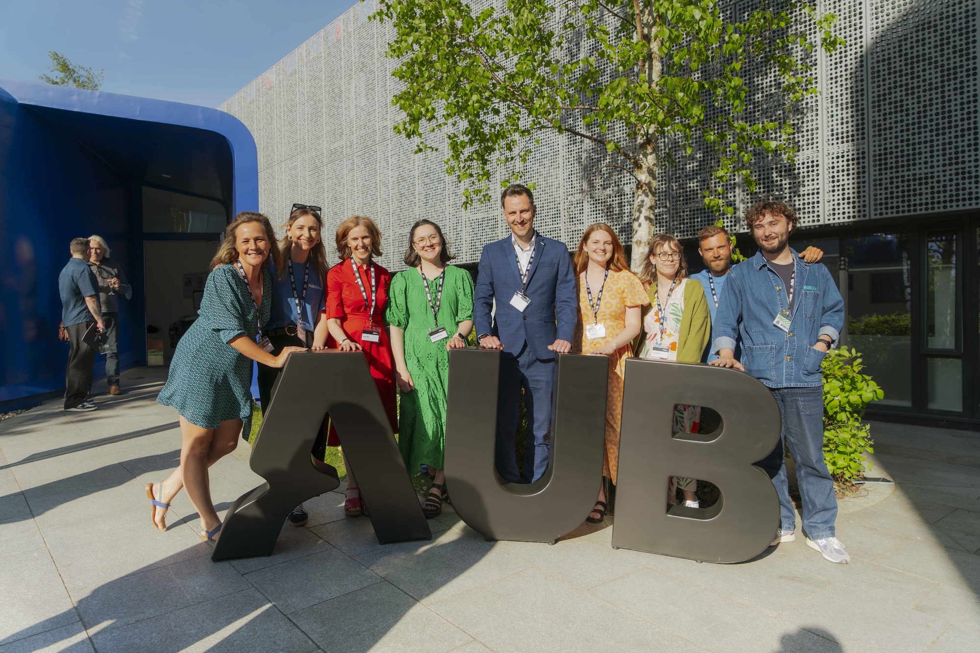 A group of nine people stands behind large letters spelling AUB outside a modern building, with a tree and blue entrance arch in the background.