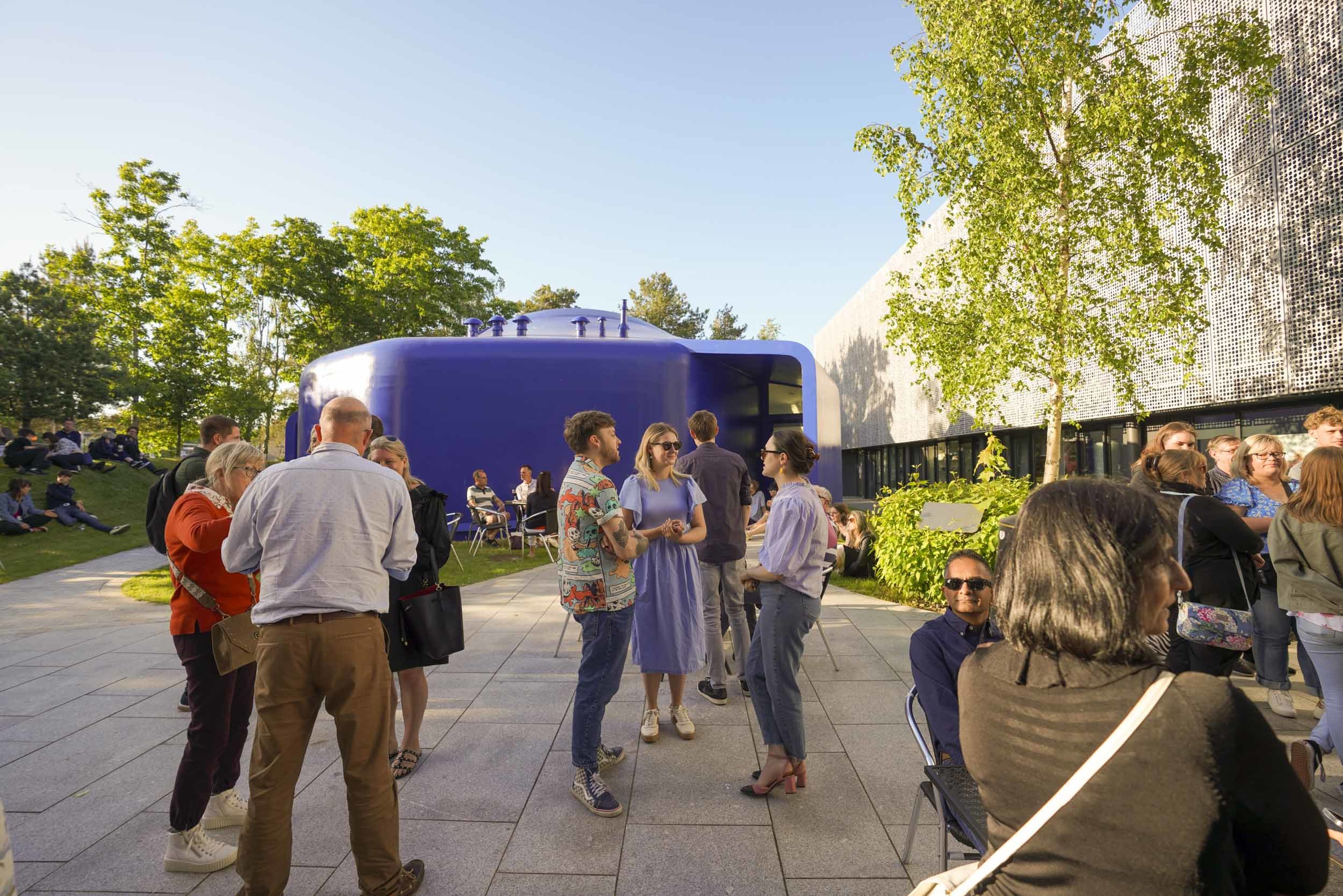 Groups of people standing in a courtyard outside a blue university building at an event.