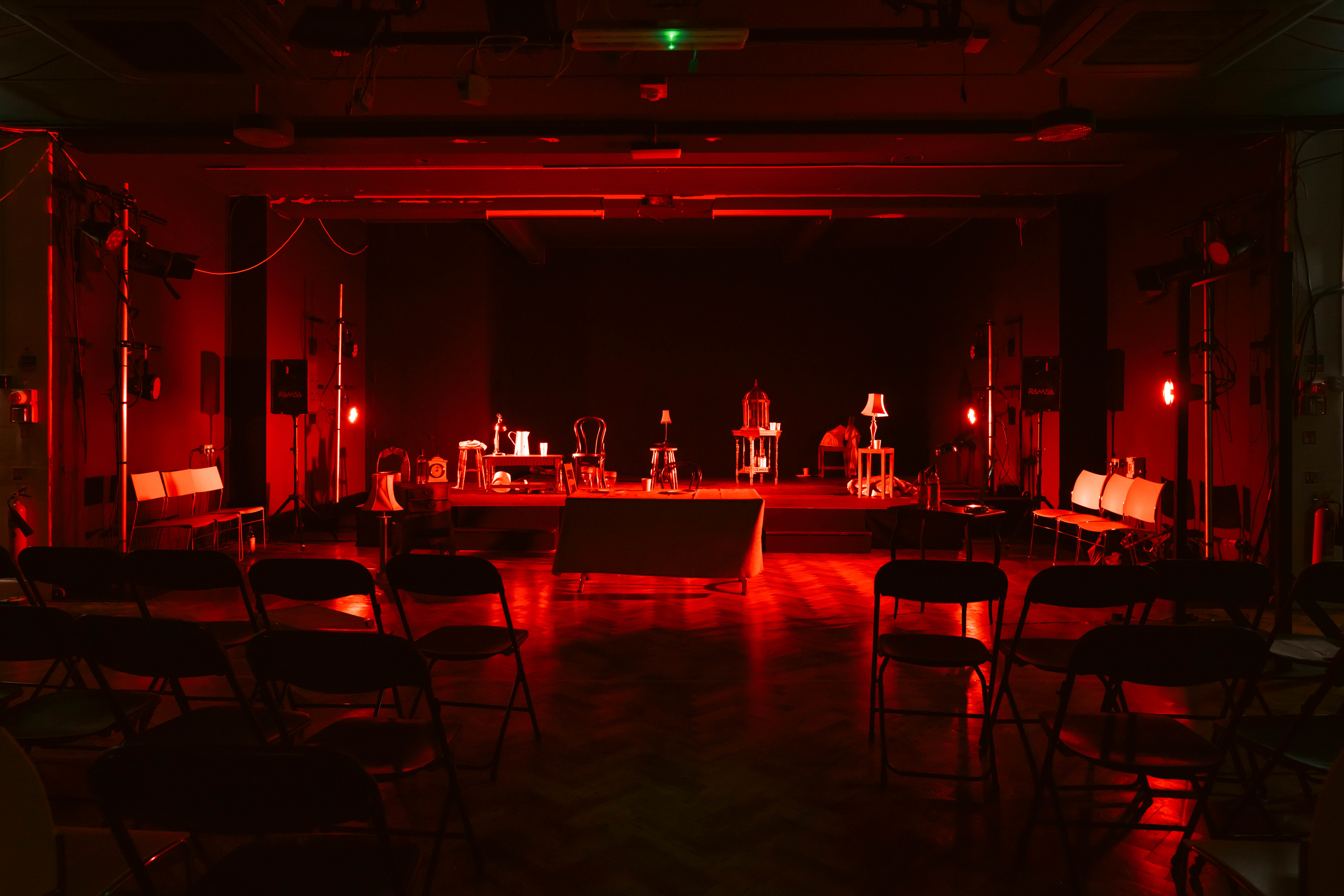 A stage set with miscellaneous pieces of furniture with heavy red lighting. Empty seats are visible in the foreground.