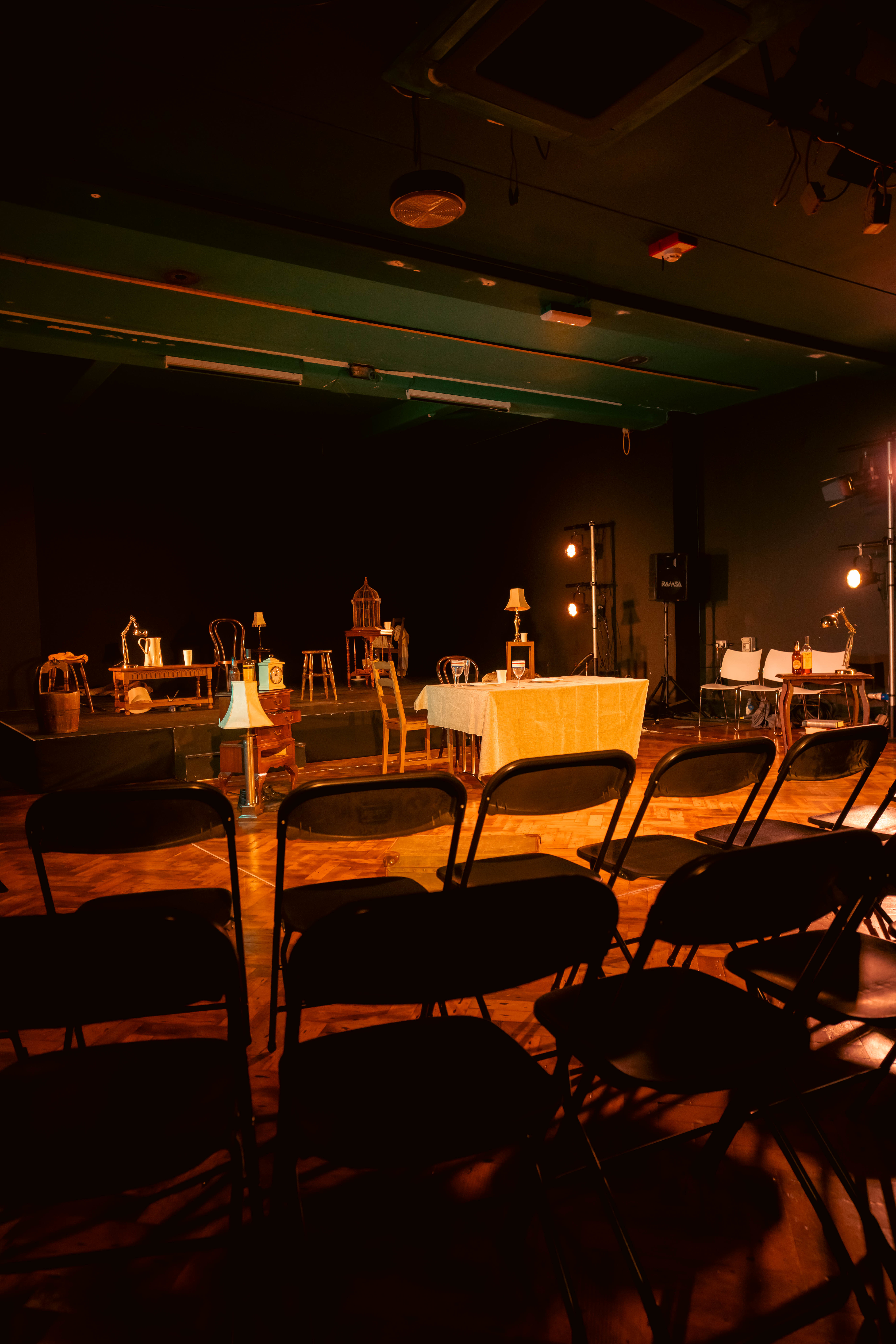 A stage set with assorted pieces of furniture including tables, chairs, stools, a lamp and a birdcage. Rows of empty seats appear in the foreground.