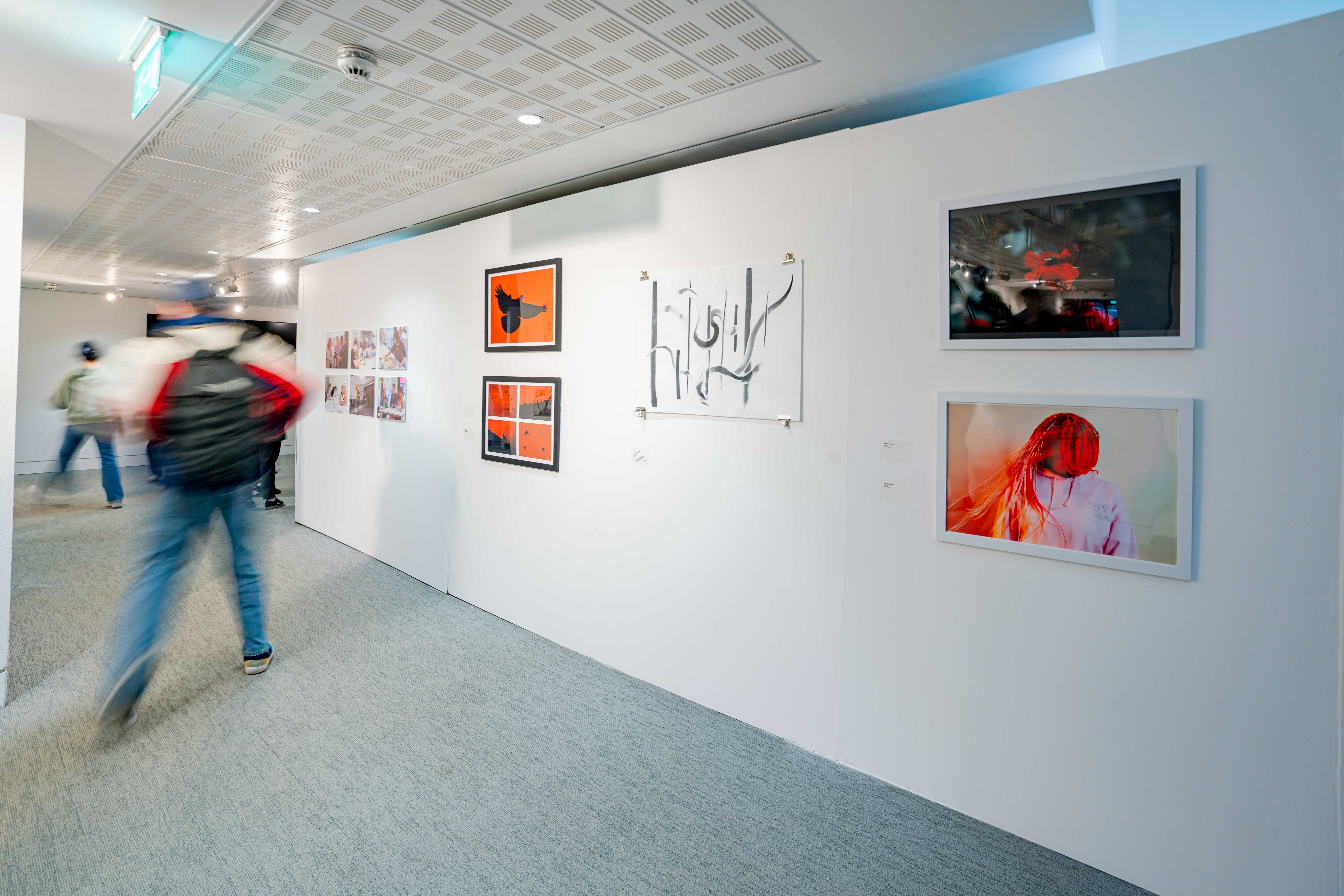 Image of a wall in an art gallery displaying four large red coloured photographs and a black and white large ink drawing