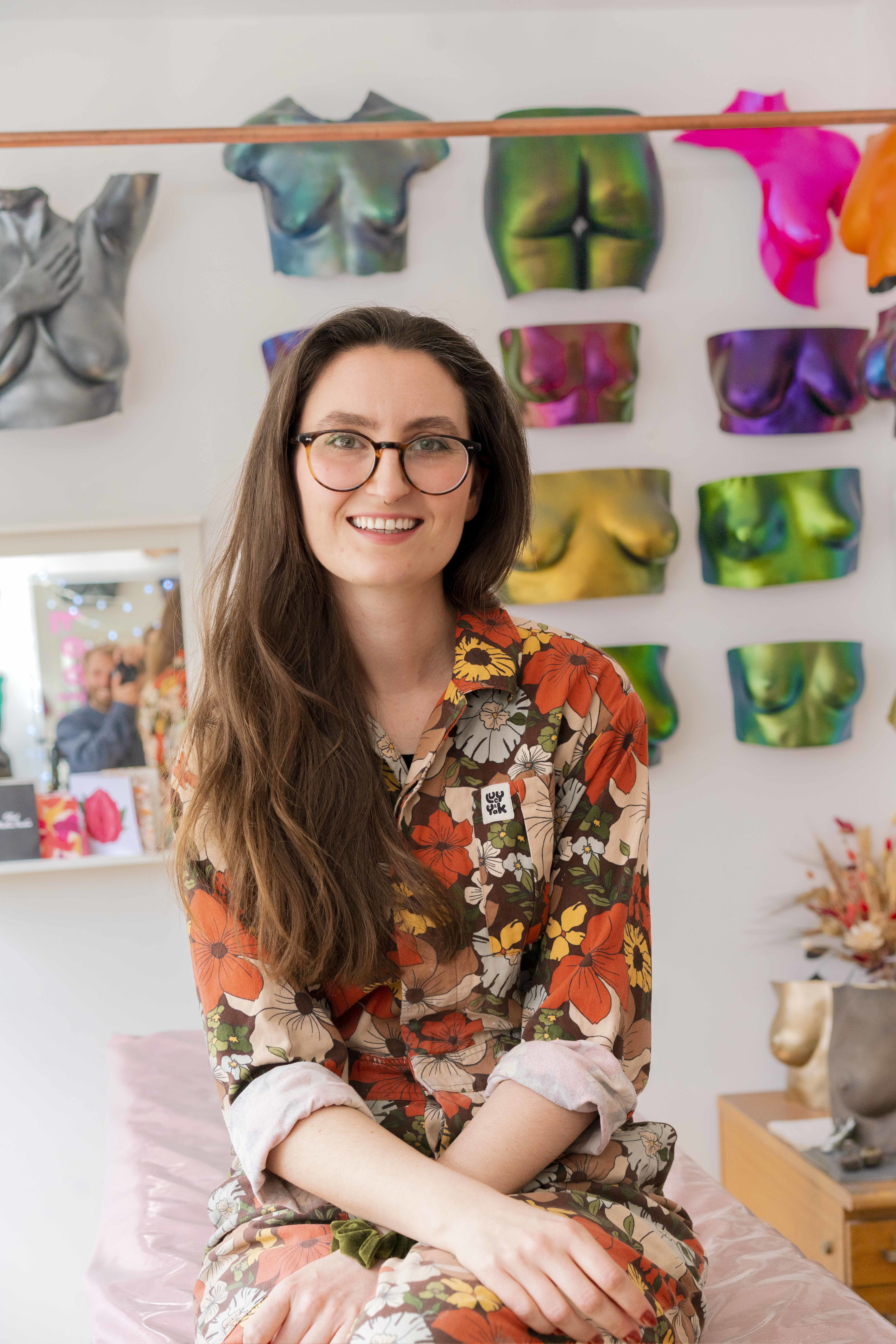 Lydia Reeves sat in her studio smiling, with colourful, glittery castings of vulvas hung on the wall behind her.