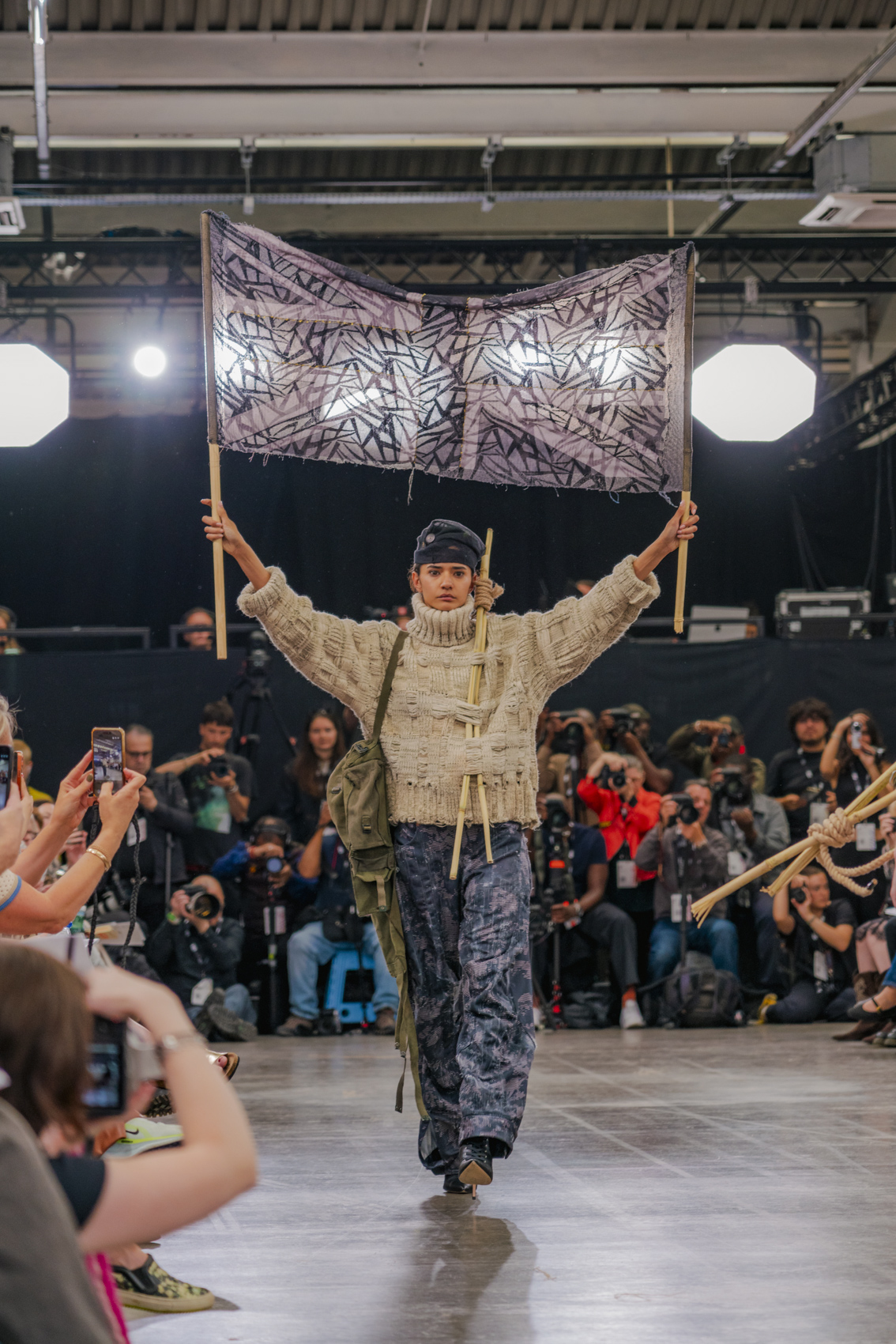 Model walking down catwalk holding a large, purple triangle Union Jack flag, threaded beige top and blue trousers.