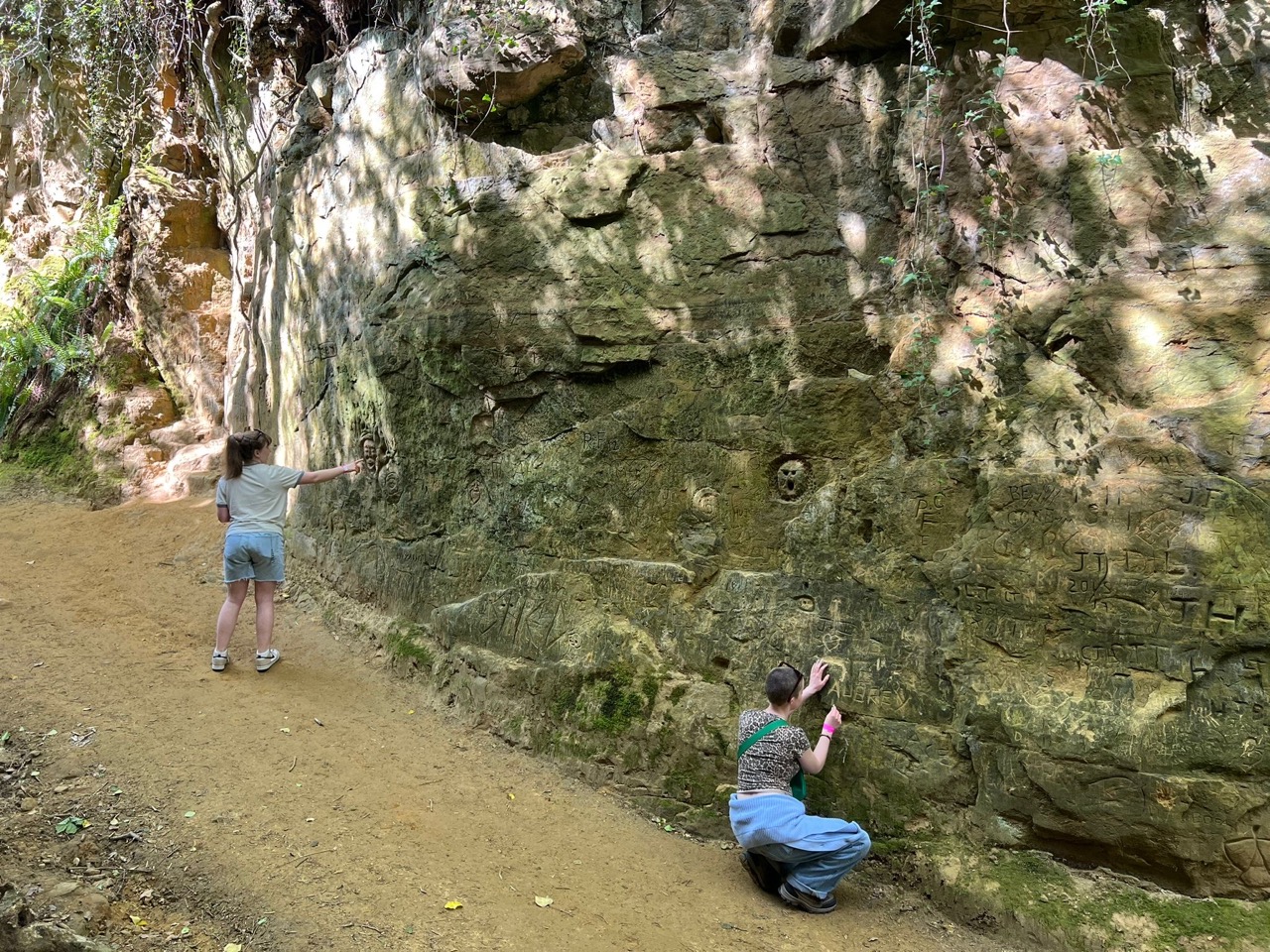 Two people stood by a tall, mossy rock wall with inscriptions and etchings, the person on the right etching 'AUB' onto them.