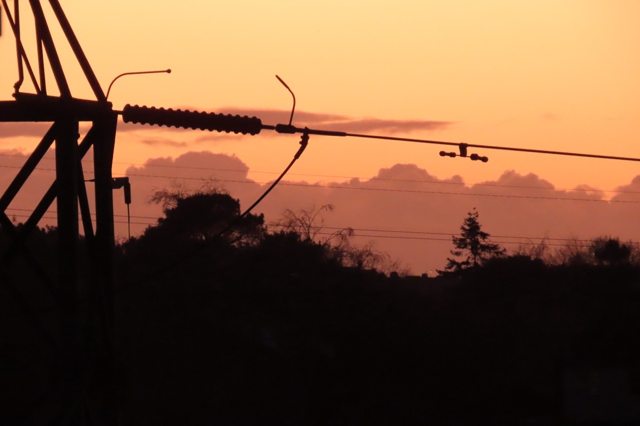 Power lines and electrical infrastructure silhouetted against orange sunset sky with dark trees and purple-tinged clouds.