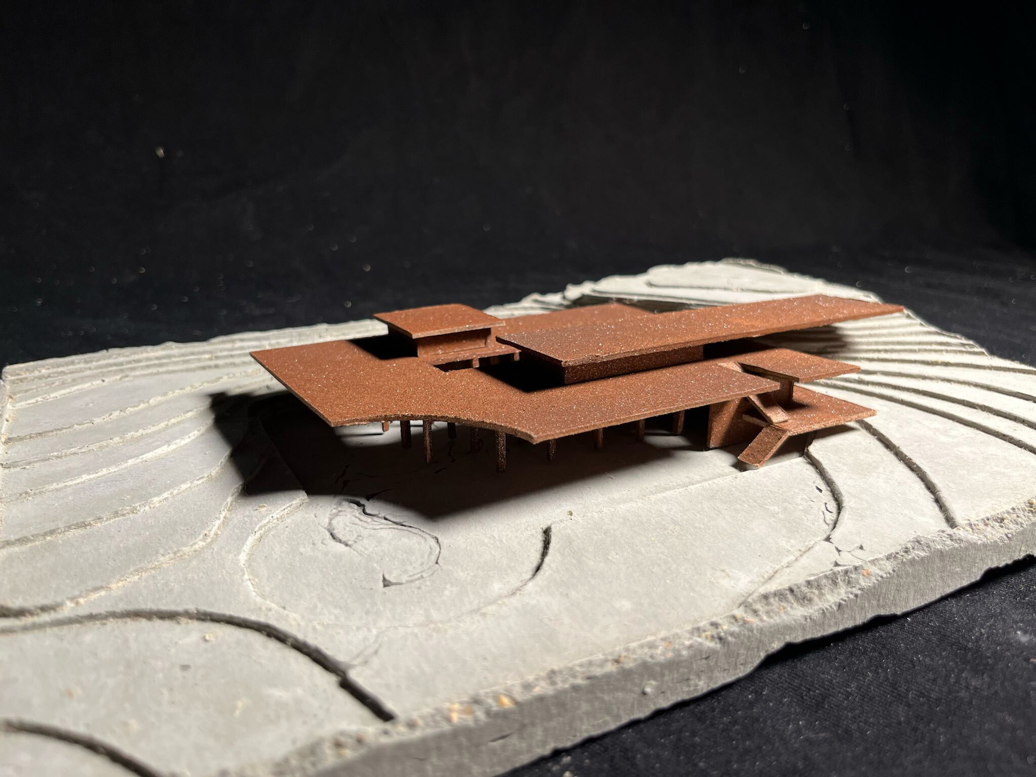 Architectural model with brown rectangular buildings on curved white base, casting shadows in bright sunlight against dark background.