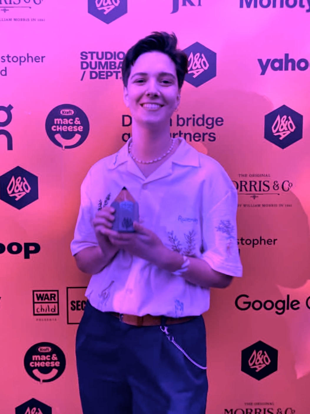 Young person smiling whilst holding award trophy at event, standing before pink backdrop with multiple sponsor logos including Yahoo, Google, and D&AD.
