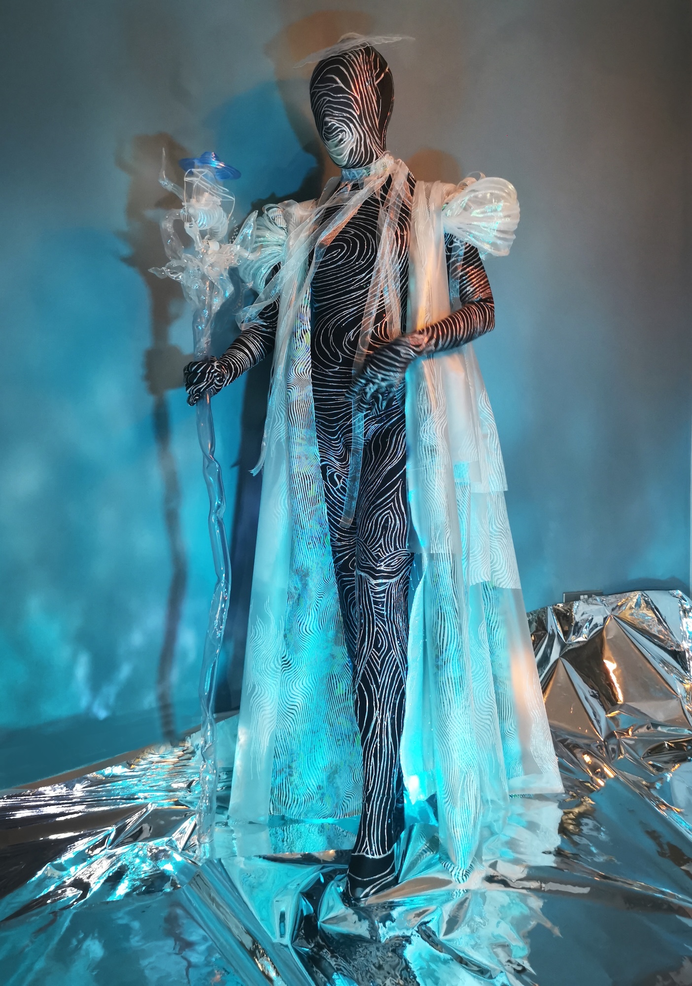 Figure in black bodysuit with white linear patterns, draped in translucent white fabric, standing amongst reflective materials under blue lighting.