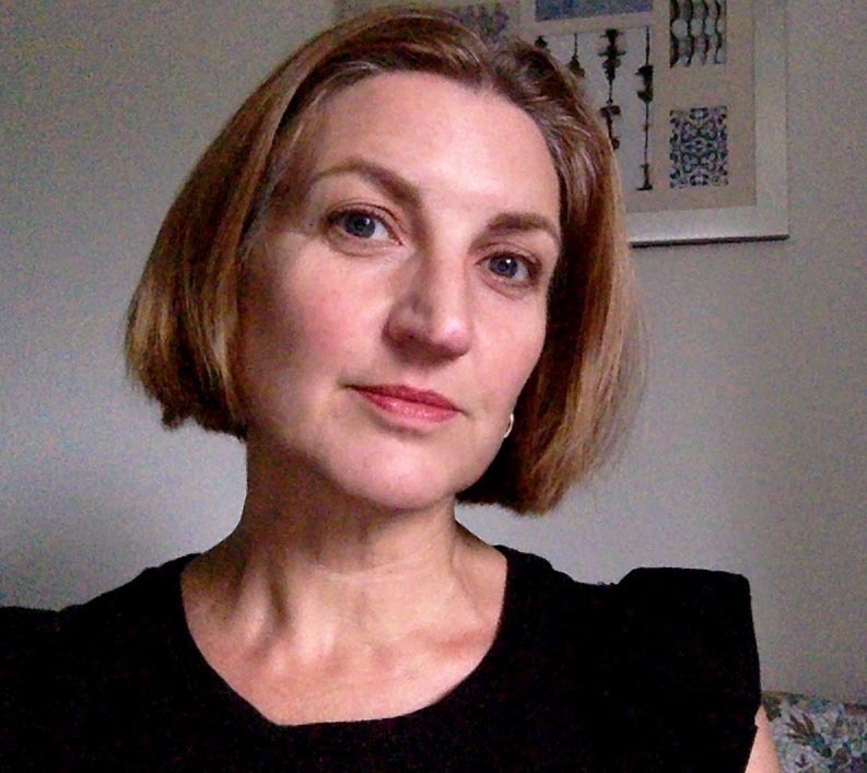 Woman with shoulder-length brown hair wearing black top, looking directly at camera. White wall with framed artwork visible behind.