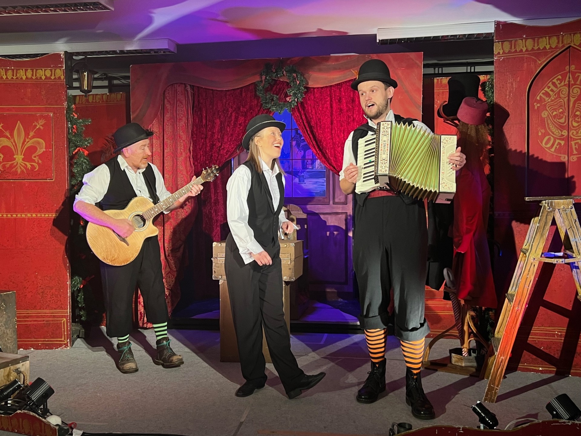 Three performers wearing white shirts and black trousers and waistcoats performing A Christmas Carol. They are all singing. One is playing the guitar, another is playing an accordion.