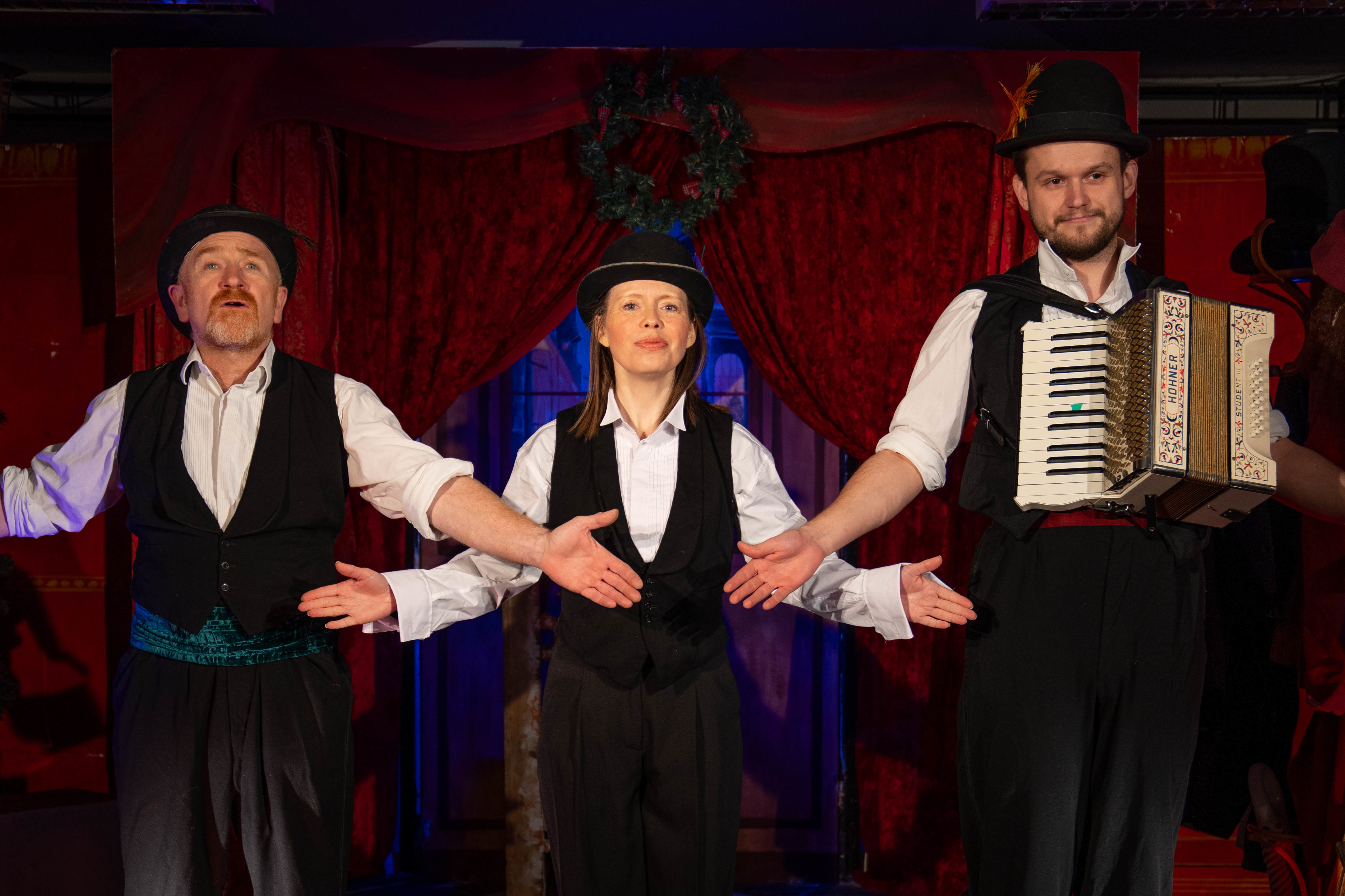 Three actors in white shirts and black waistcoats performing towards the camera. One of them is wearing a Tyrolean hat and an accordion.