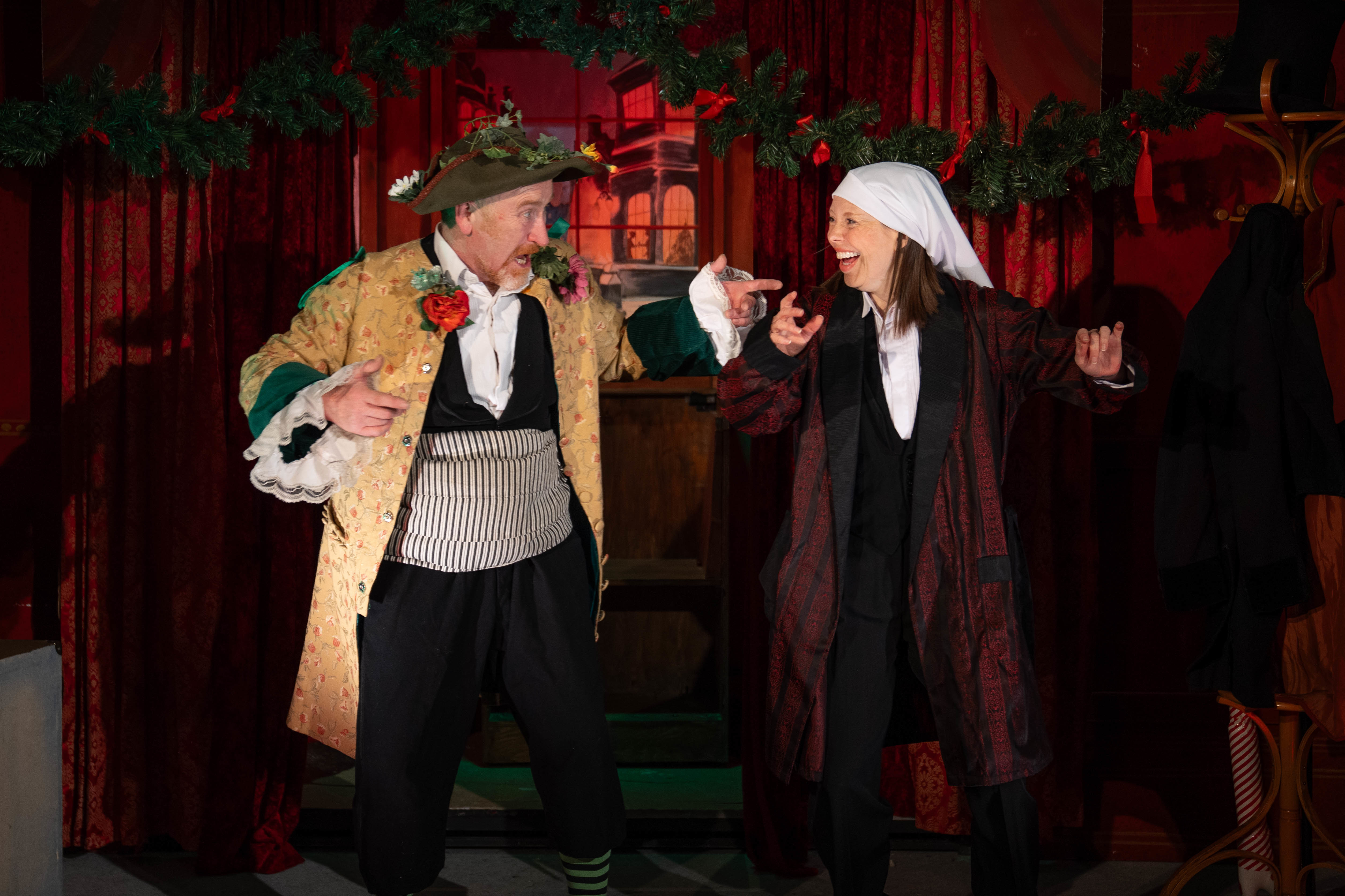 Actors in costume as Ebenezer Scrooge and the Ghost of Christmas Present interacting on a Victorian-decorated stage.