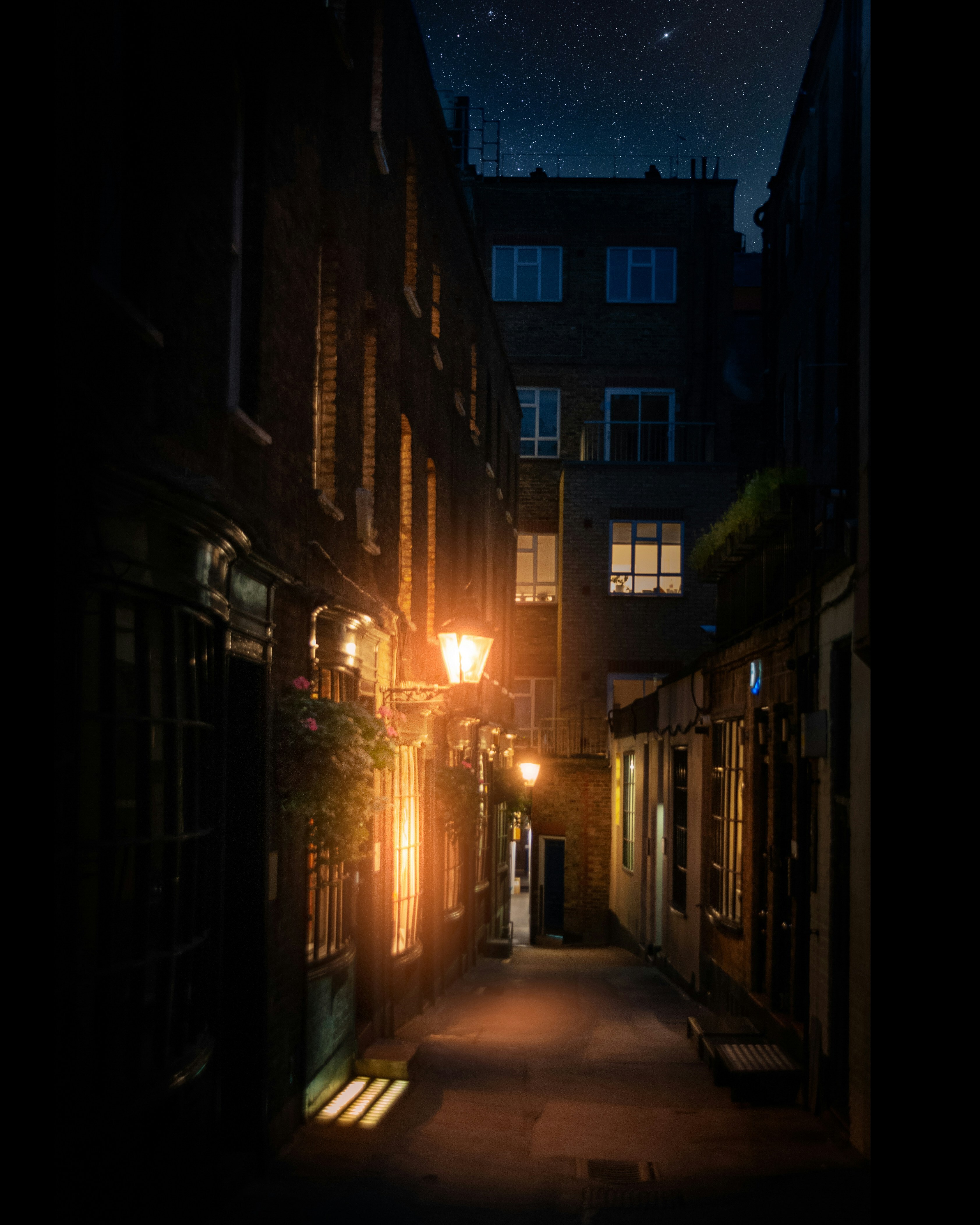 Narrow cobblestone alley at night with warm yellow streetlamp glow, dark building silhouettes, and starry blue sky above.
