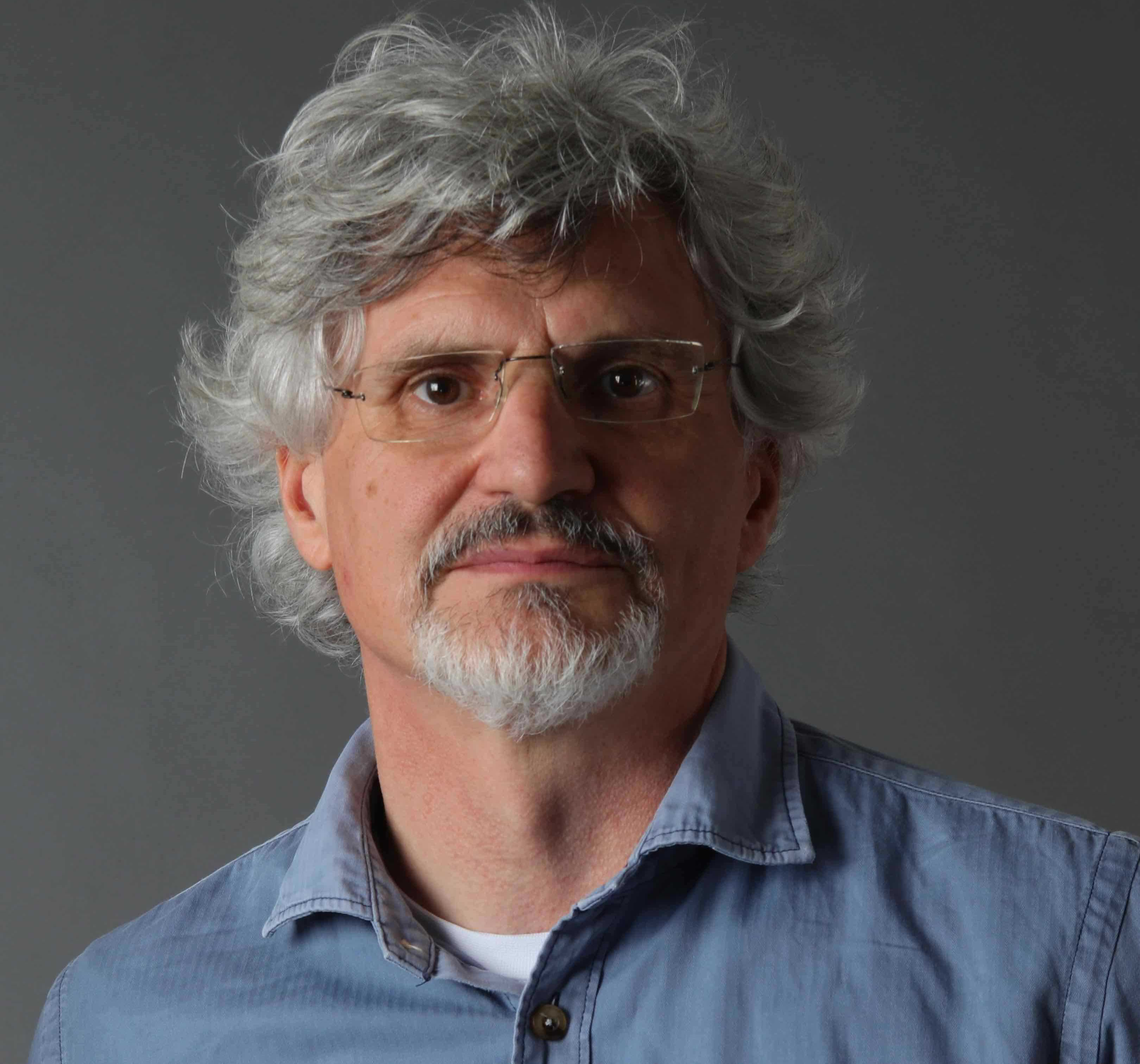Middle-aged man with grey curly hair, beard, and wire-rimmed glasses wearing blue button-up shirt against grey background.
