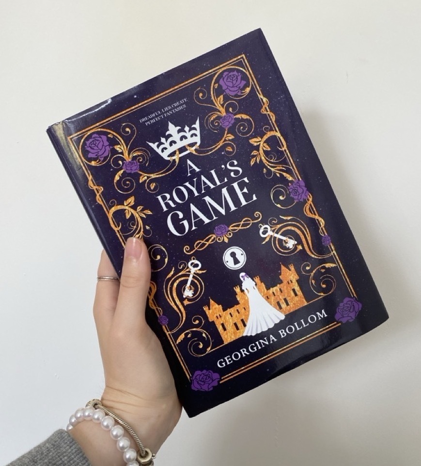 Book copy of 'A Royal's Game' with an illustrated purple and gold cover, held up against a white background.
