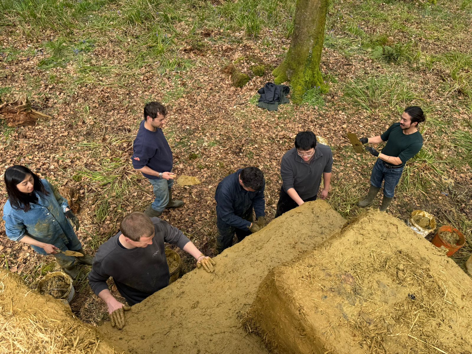 Five people working together to pile earth into a large mound in a wooded area, with buckets and leaf-covered ground nearby.