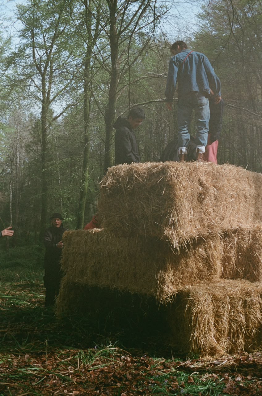A man in a blue jacket stands on stacked hay bales in a wooded clearing, with two other people nearby.
