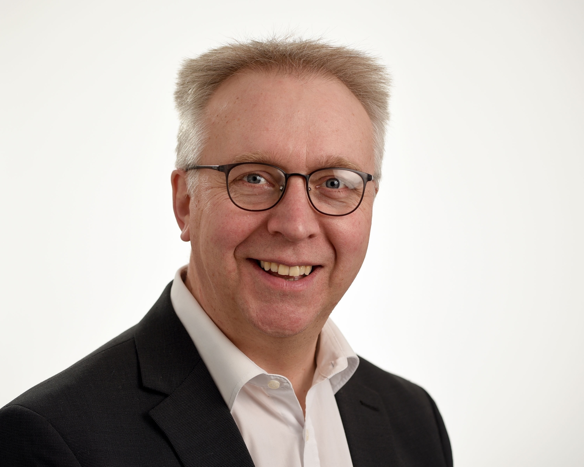 Middle-aged man with grey hair and round glasses wearing dark blazer over white shirt, smiling at camera against plain background.