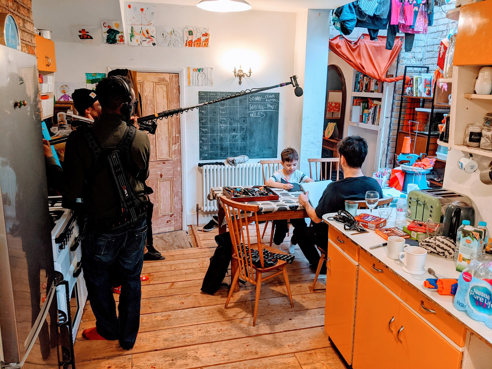 Behind the scenes of a film crew filming in a kitchen. A child and an adult are sat at a dining table painting