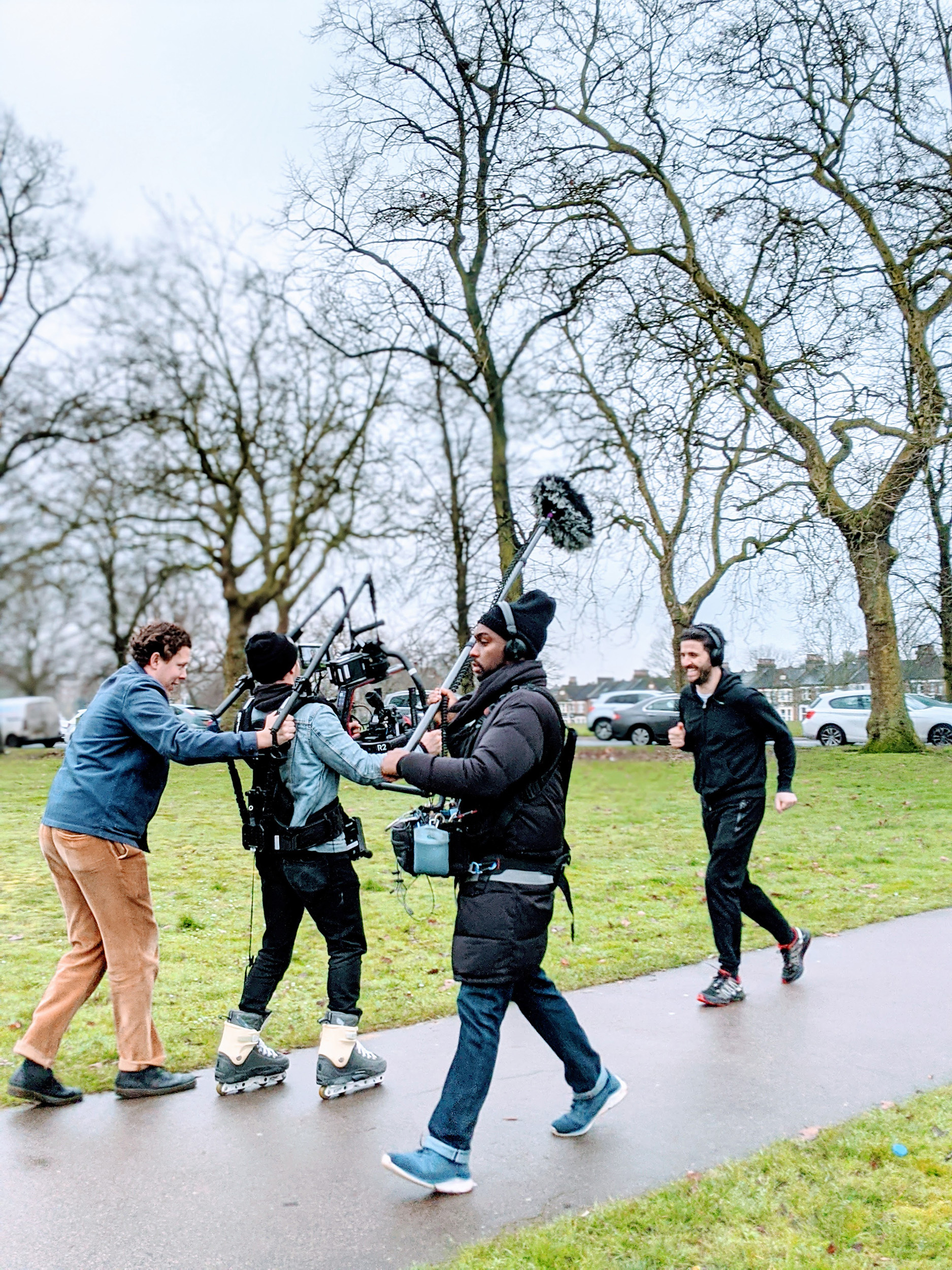 Behind the scenes of a film crew following a person jogging. One of the three members of the film crew is holding a camera while on roller skates