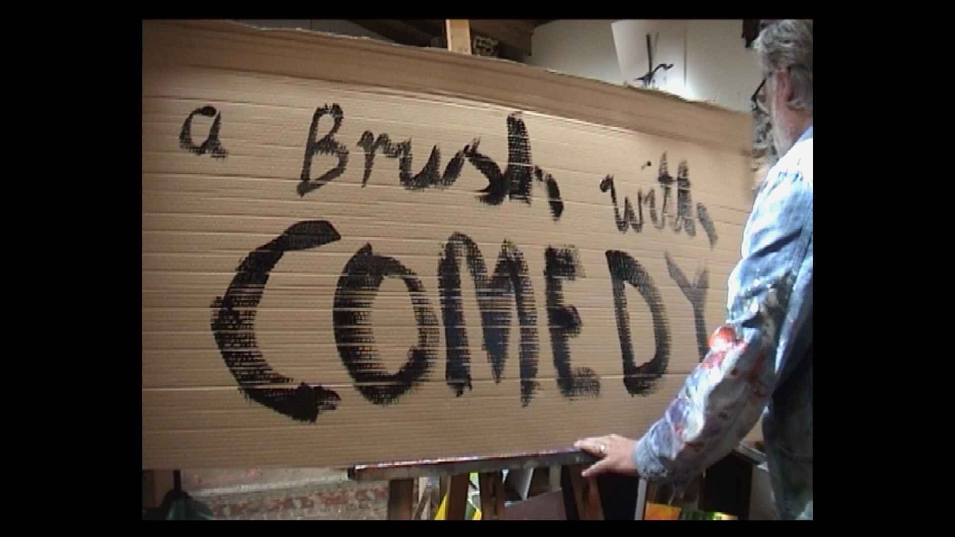 Comedian Vic Reeves painting the words 'a brush with comedy' on to a piece of cardboard with black paint
