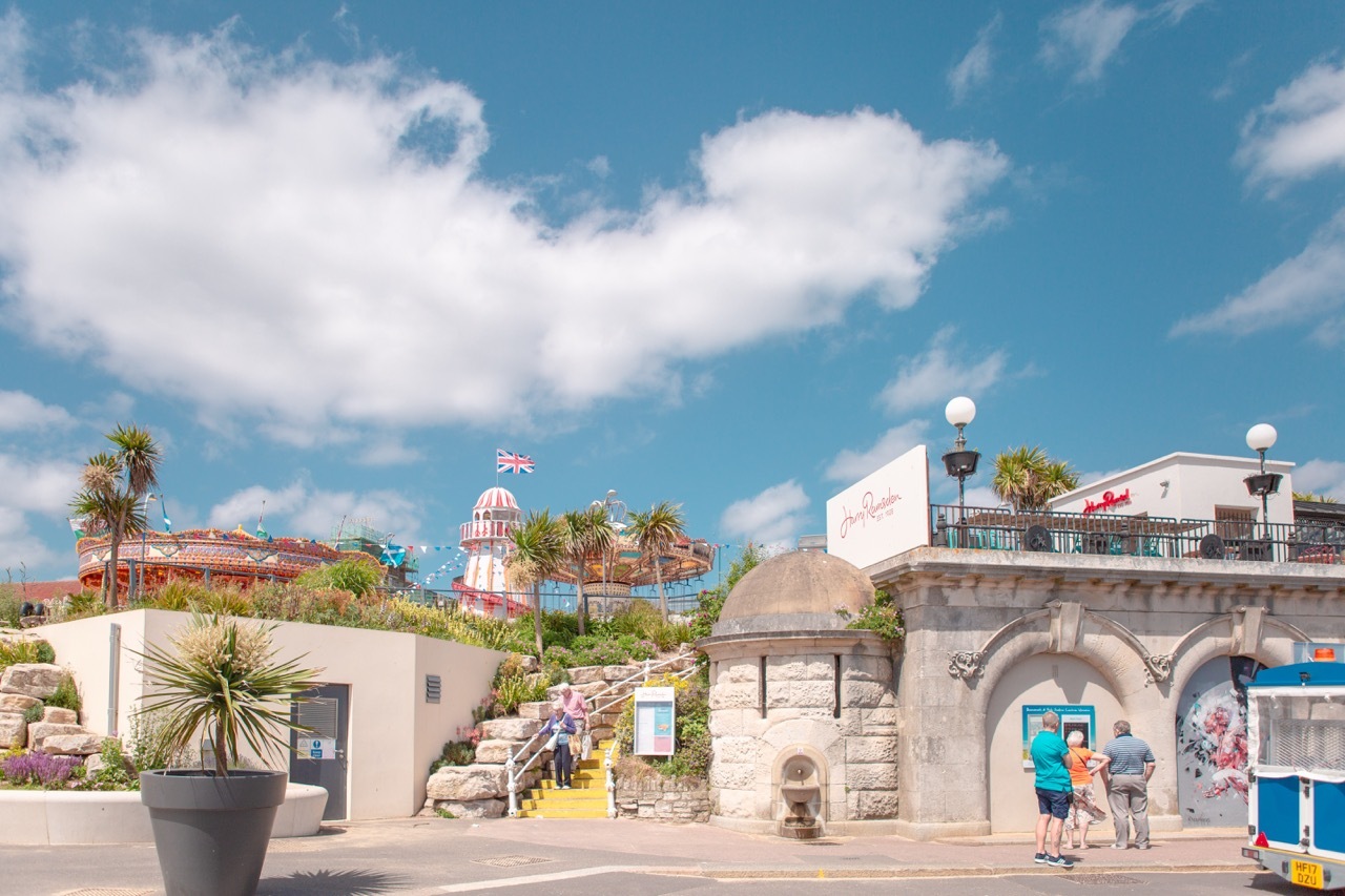 Seaside town with white and pink buildings, palm trees, blue sky with white clouds, stone archway, and people walking along waterfront promenade.