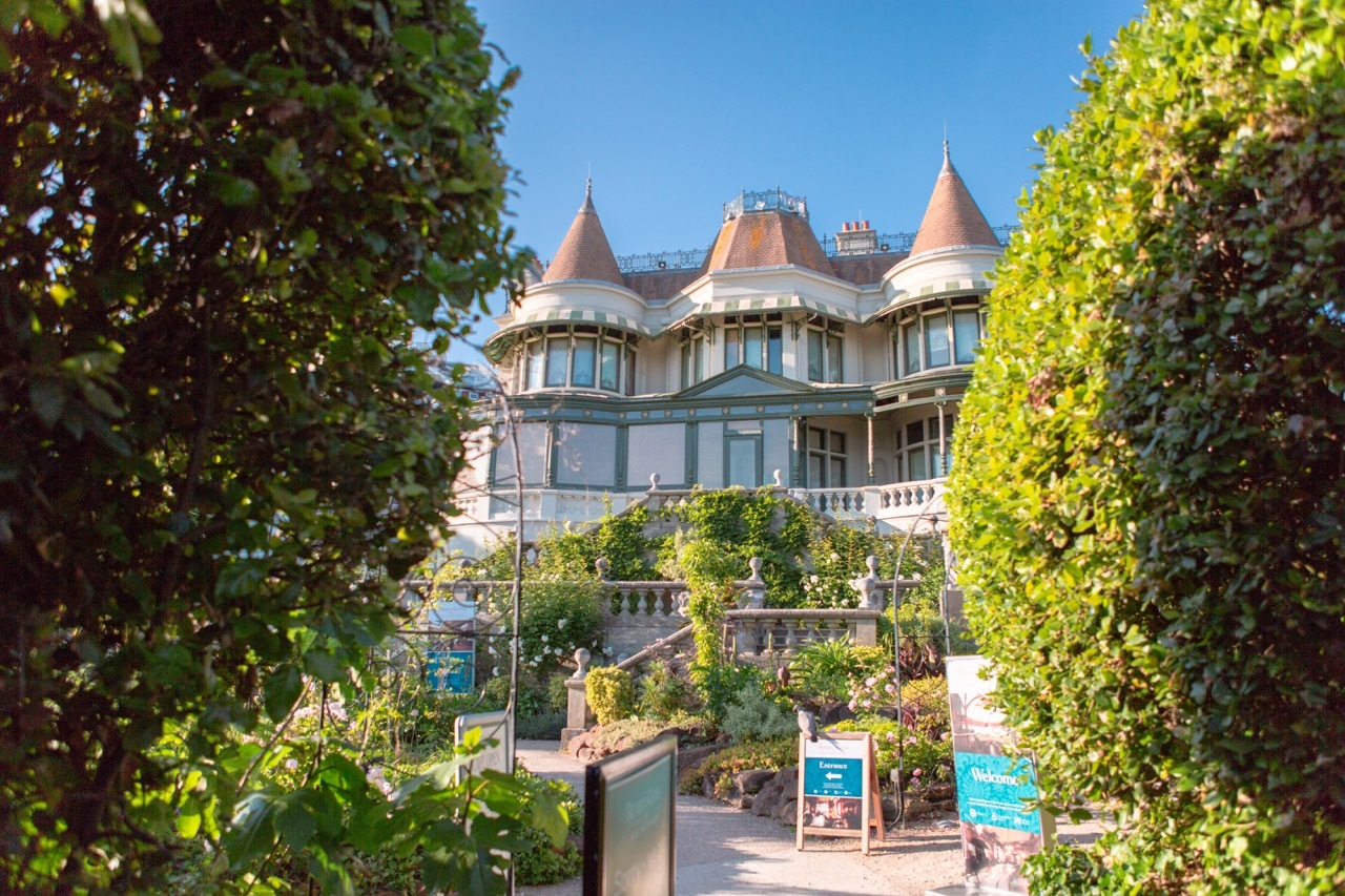 Victorian mansion with conical turrets and grey stone facade, framed by lush green trees under blue sky, with garden entrance visible.