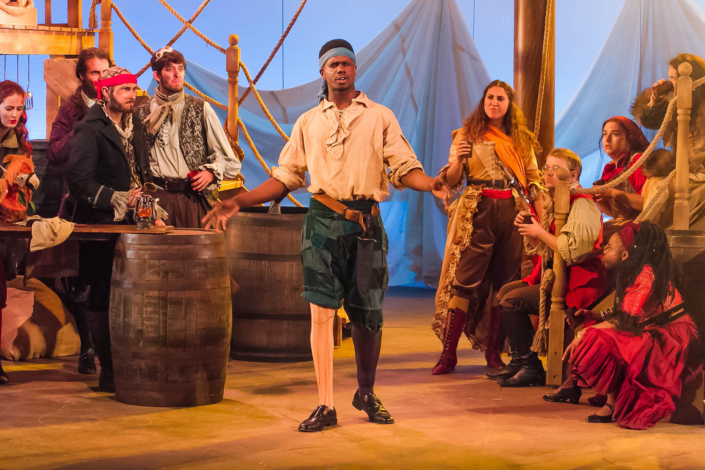 Actor in costume as Long John Silver walks centre stage surrounded by other cast members, also in costume as pirates, sitting around barrels and on steps.