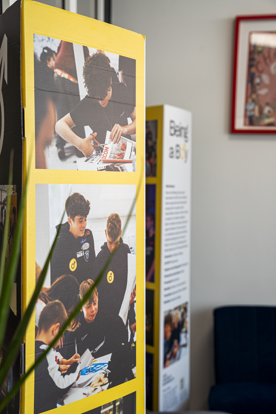 Two display panels in a room. One features a collage of photographs showing young boys engaged in various activities, including writing and drawing. The second panel presents text about the theme 'Being a Boy', alongside the edge of a blue chair and a green plant.