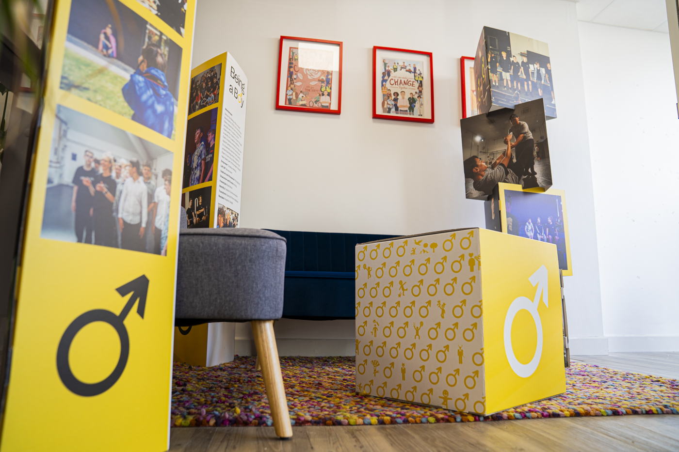 A corner of a room featuring two display boards with yellow backgrounds and male symbols, alongside a white table block with the same symbol. Red-framed images hang on the wall above a blue sofa, with a colourful rug on the wooden floor.
