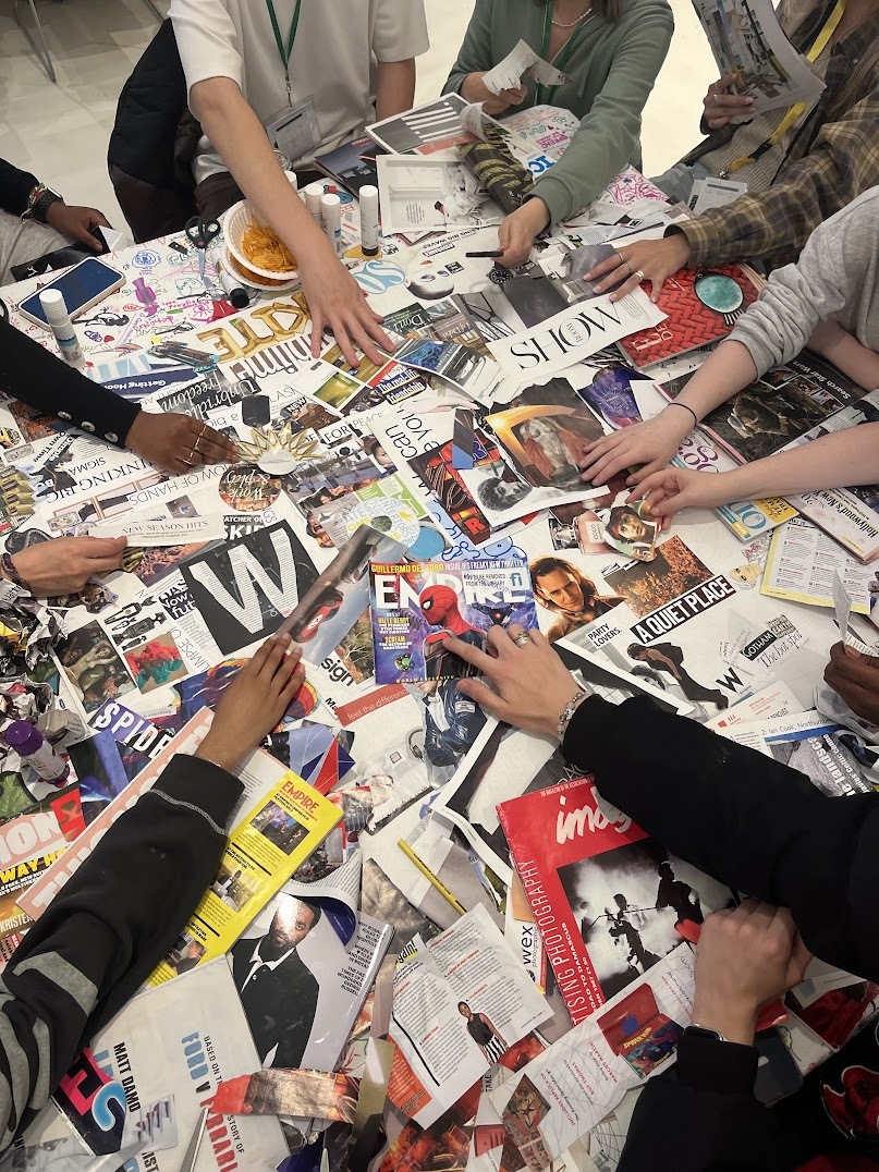 A group of student's arms around a table covered in magazines and newspaper collage