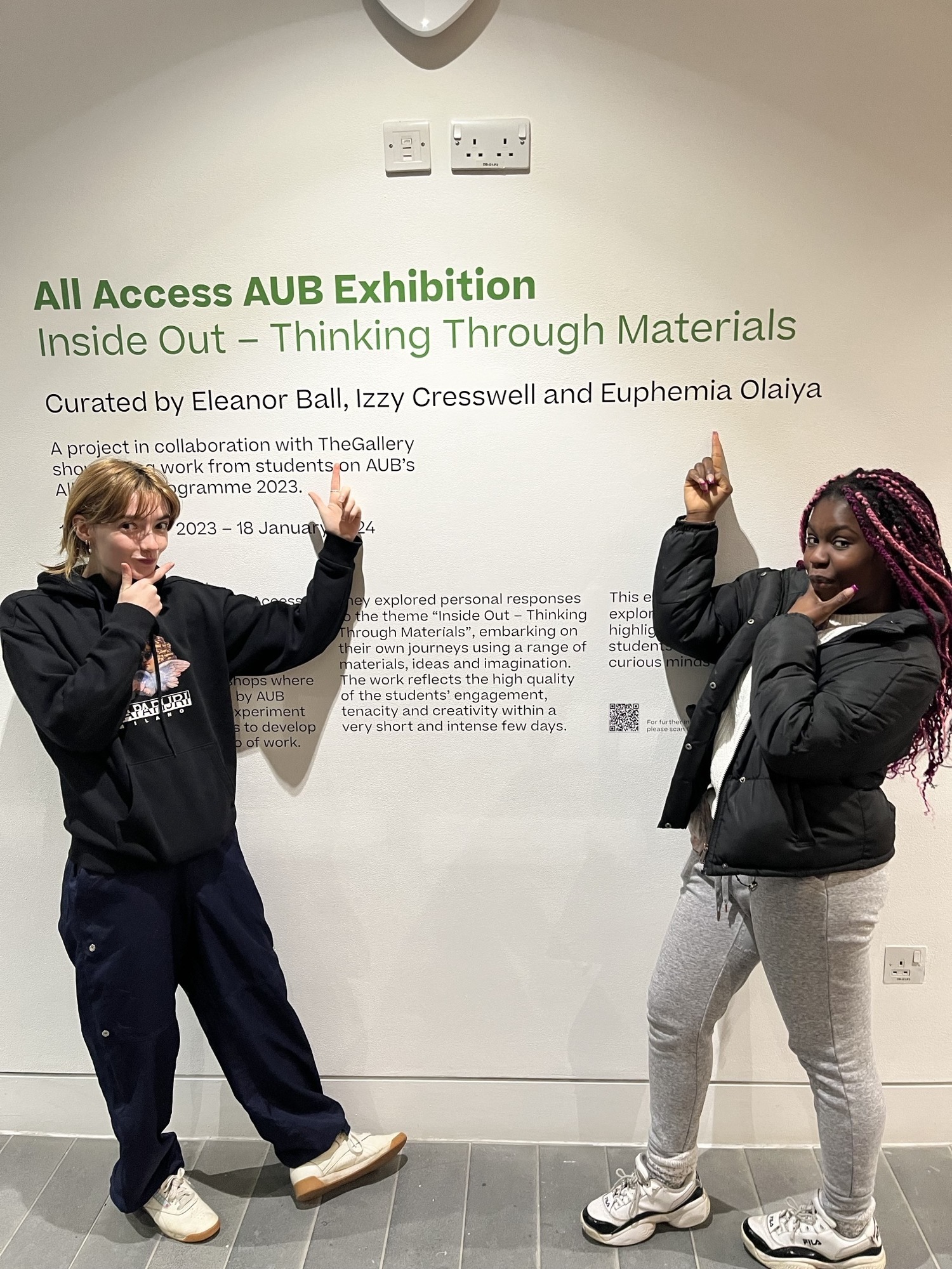 Two AUB Outreach Ambassadors posing while pointing at their names printed on a gallery wall as part of an exhibition at TheGallery, AUB.