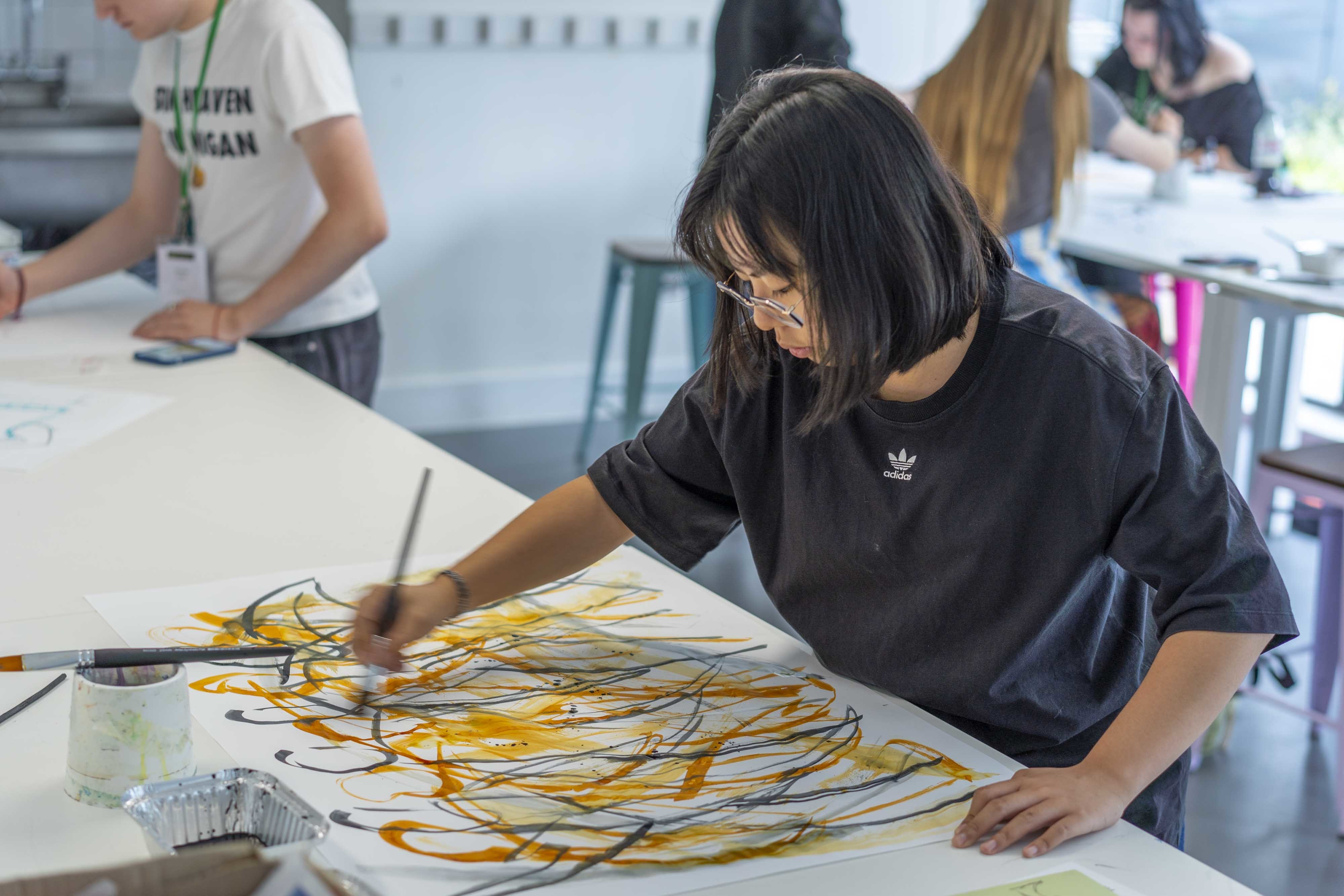 An All Access AUB student at a table painting an abstract painting in mainly yellow, orange and black strokes.