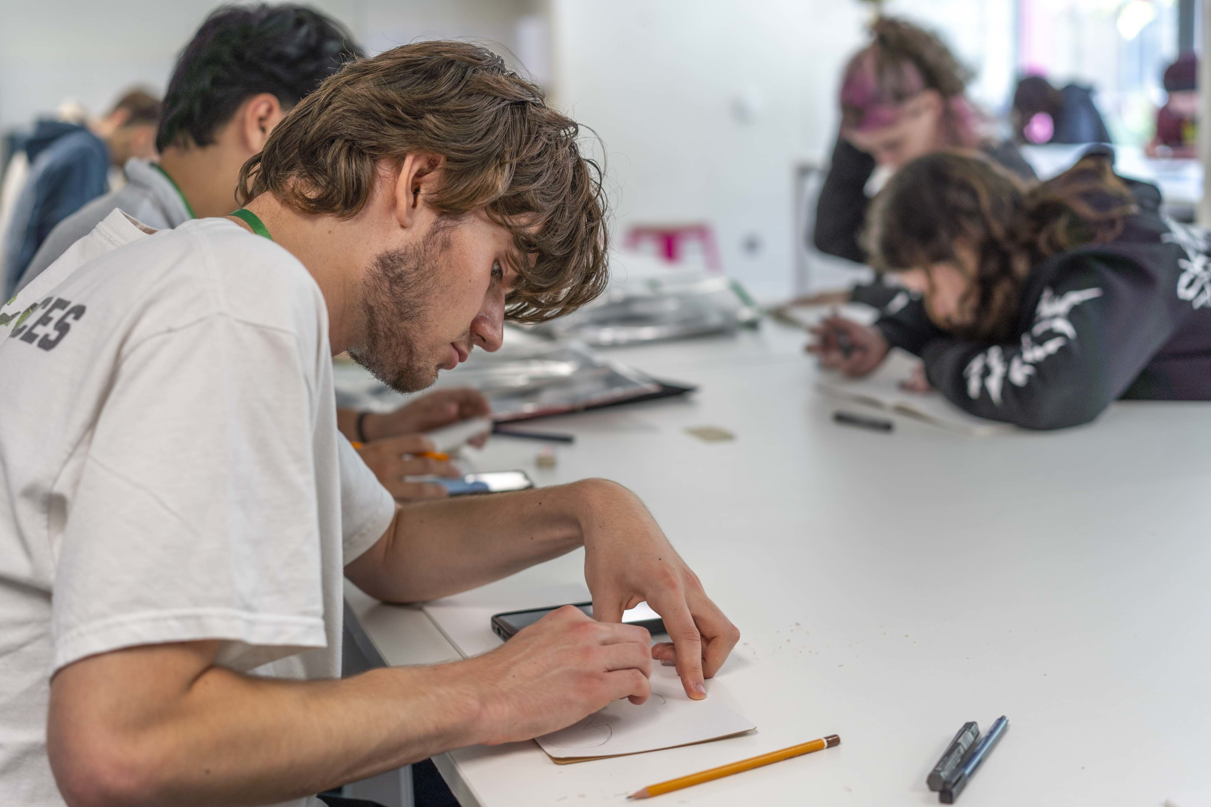 Close-up of an All Access AUB student working with a small piece of card in front of him. Fellow students are sat working around the same table.