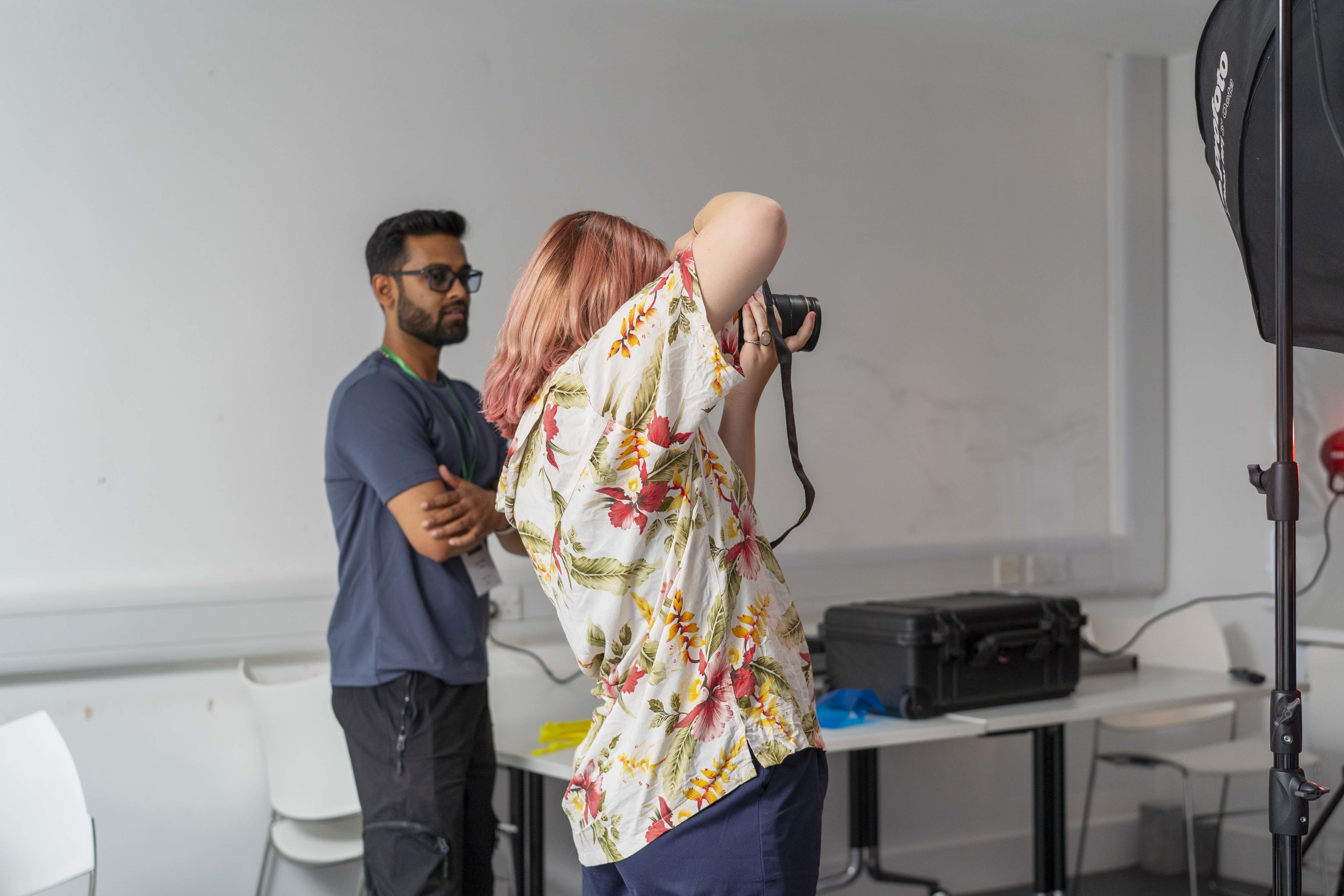 An All Access AUB student in a seminar room holding a camera portrait and looking through it, while a member of staff supervises.