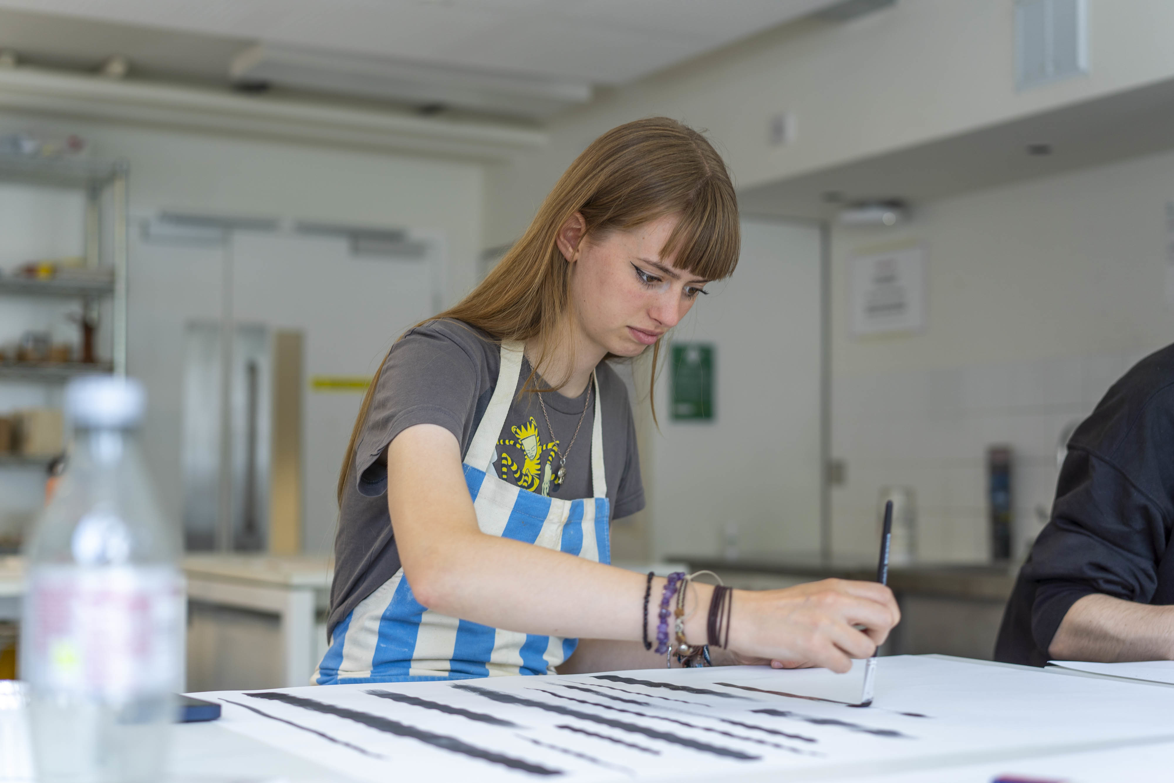 An All Access AUB student working at a table, painting in thick black vertical strokes.