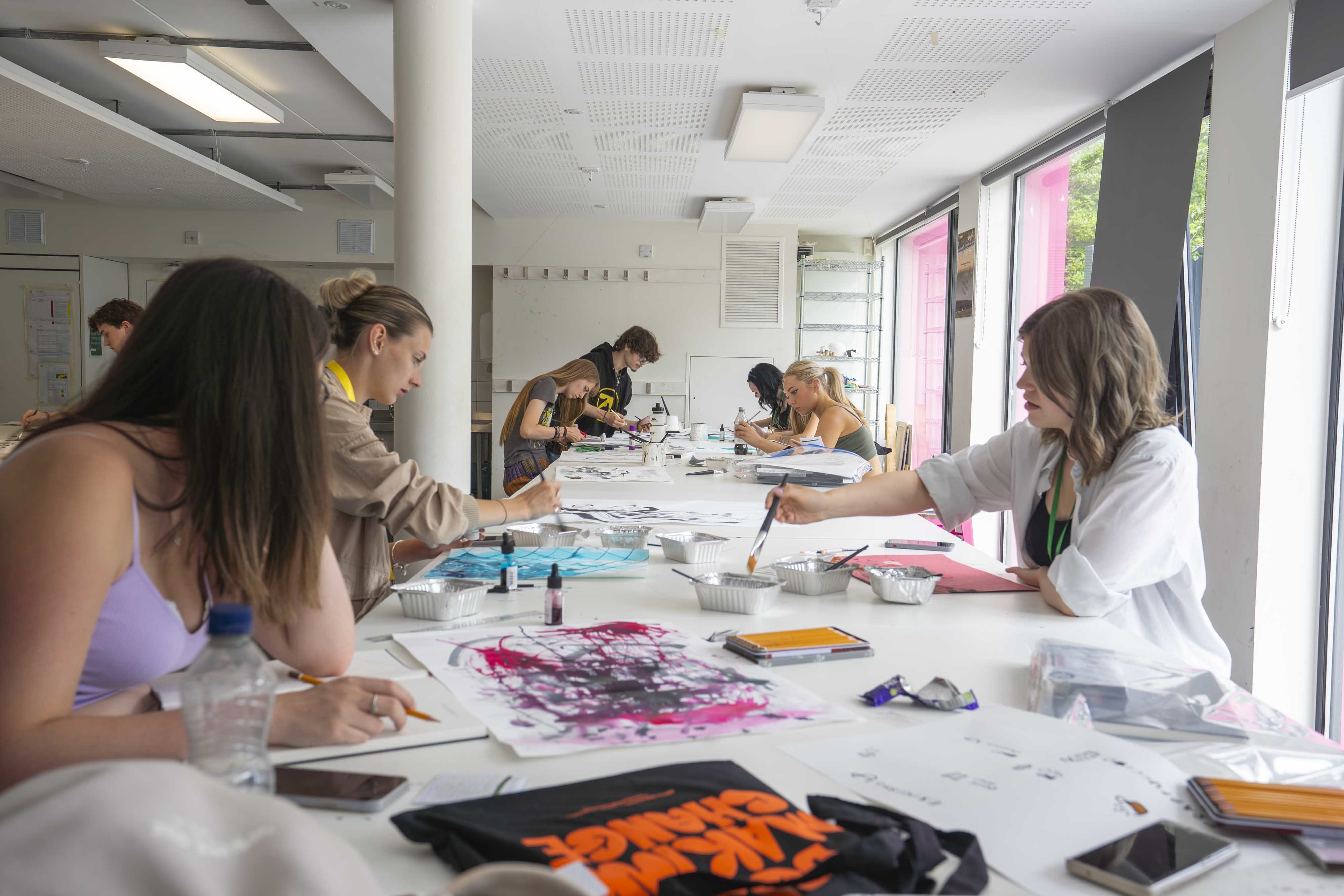 A studio with 6 students around a long table working with pink and black paint on large pieces of paper