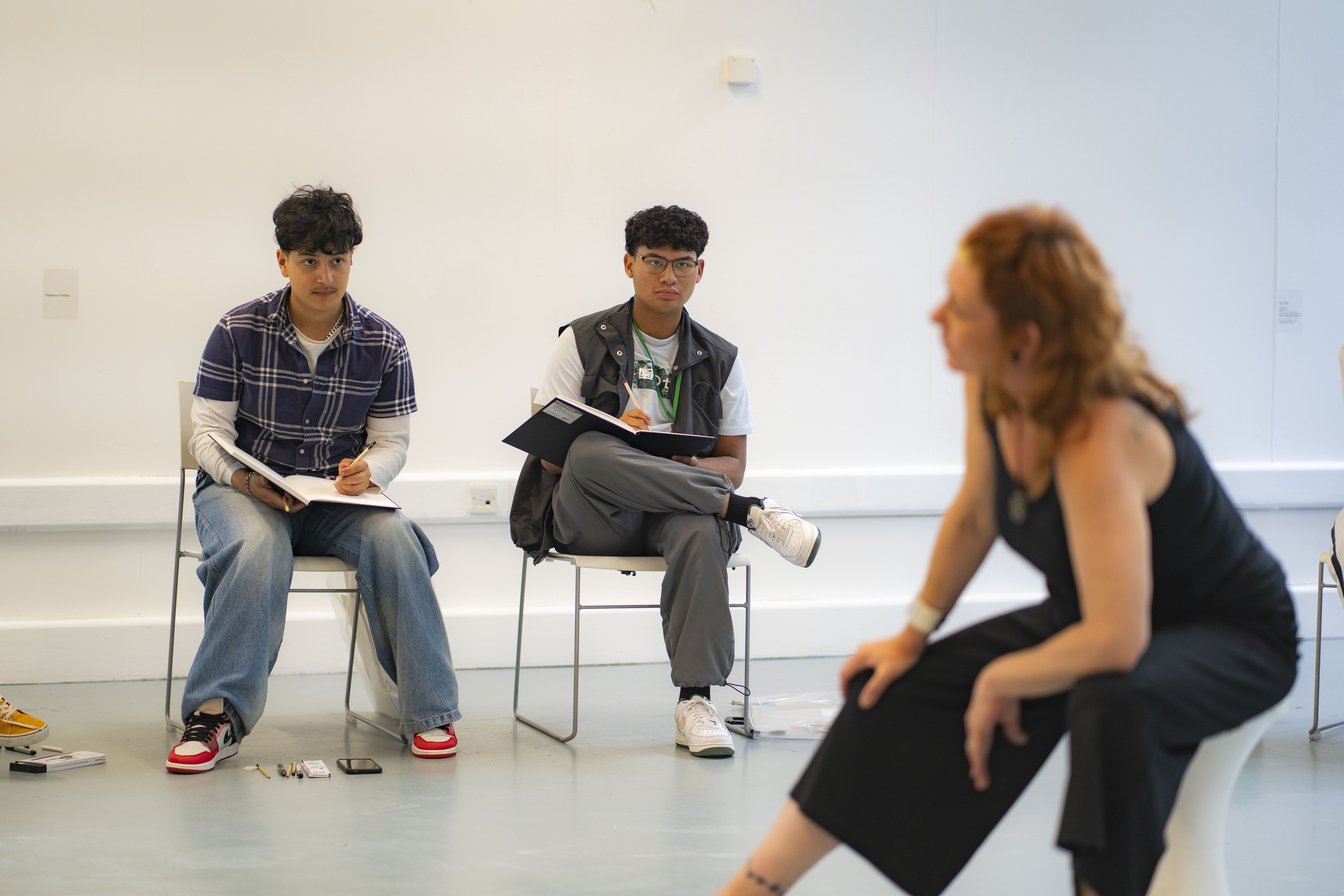 Two young people seated on white chairs in a bright, minimalist room. One is taking notes in a notebook, while the other waits beside him. In the foreground, a woman in a black outfit sits on a white stool, facing away from the camera.